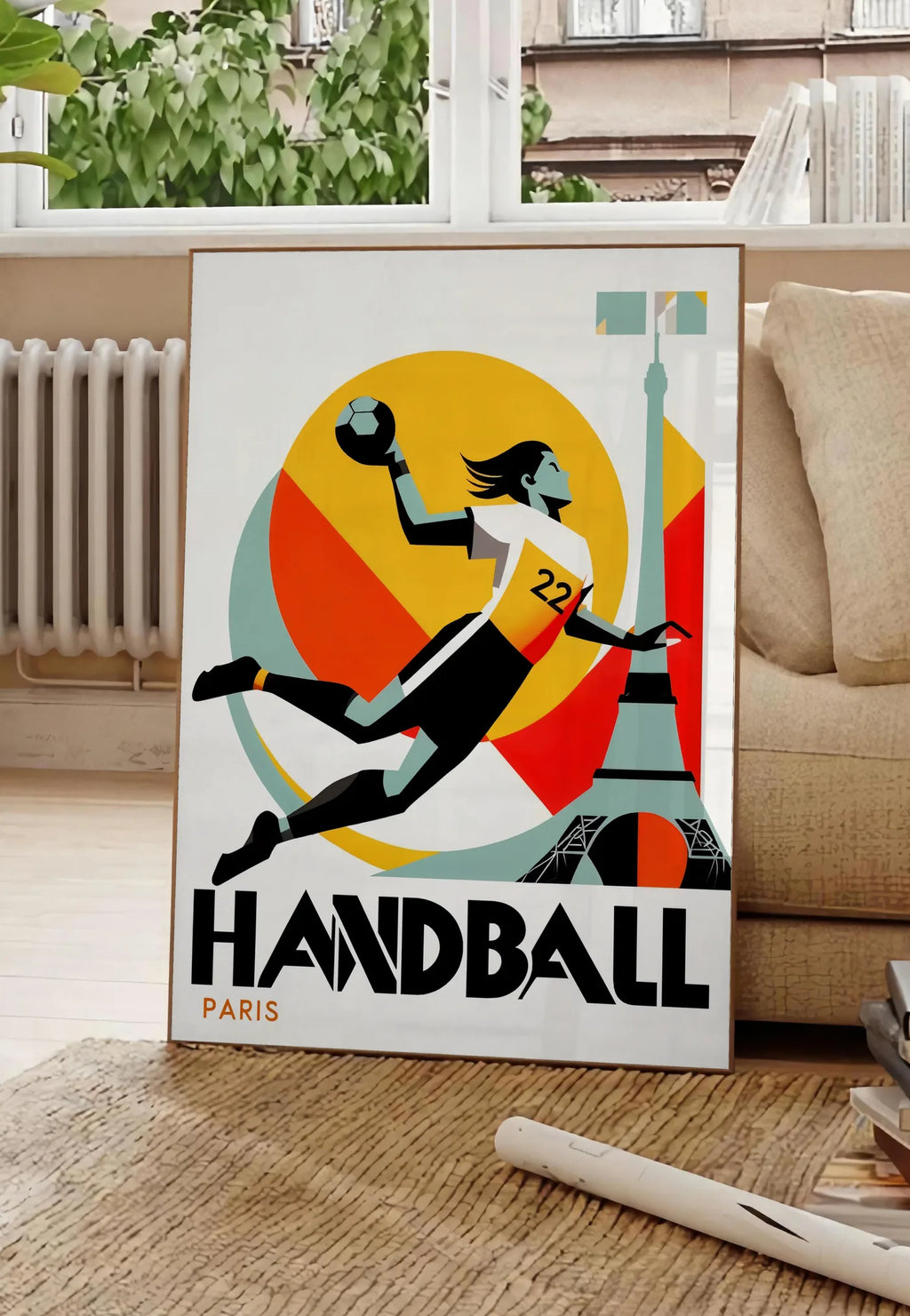 Poster affiche handball Paris, posee au sol dans un salon parisien, devant une grande fenetre donnant sur un immeuble haussmannien, entre un ficus et un canape creme