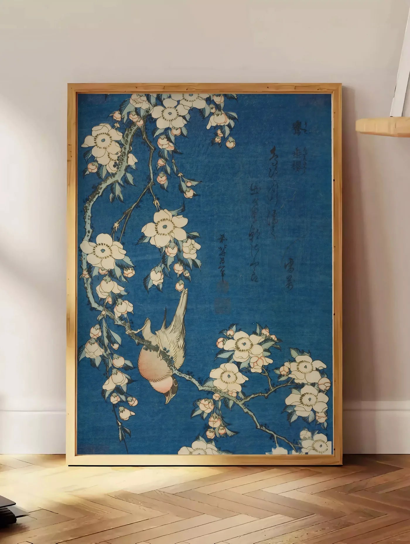 Estampe affiche Hokusai bouvreuil sur cerisier, posee au sol dans un coin salon, sur un parquet a chevrons, pres d'une table d'appoint en chene chargee de livres