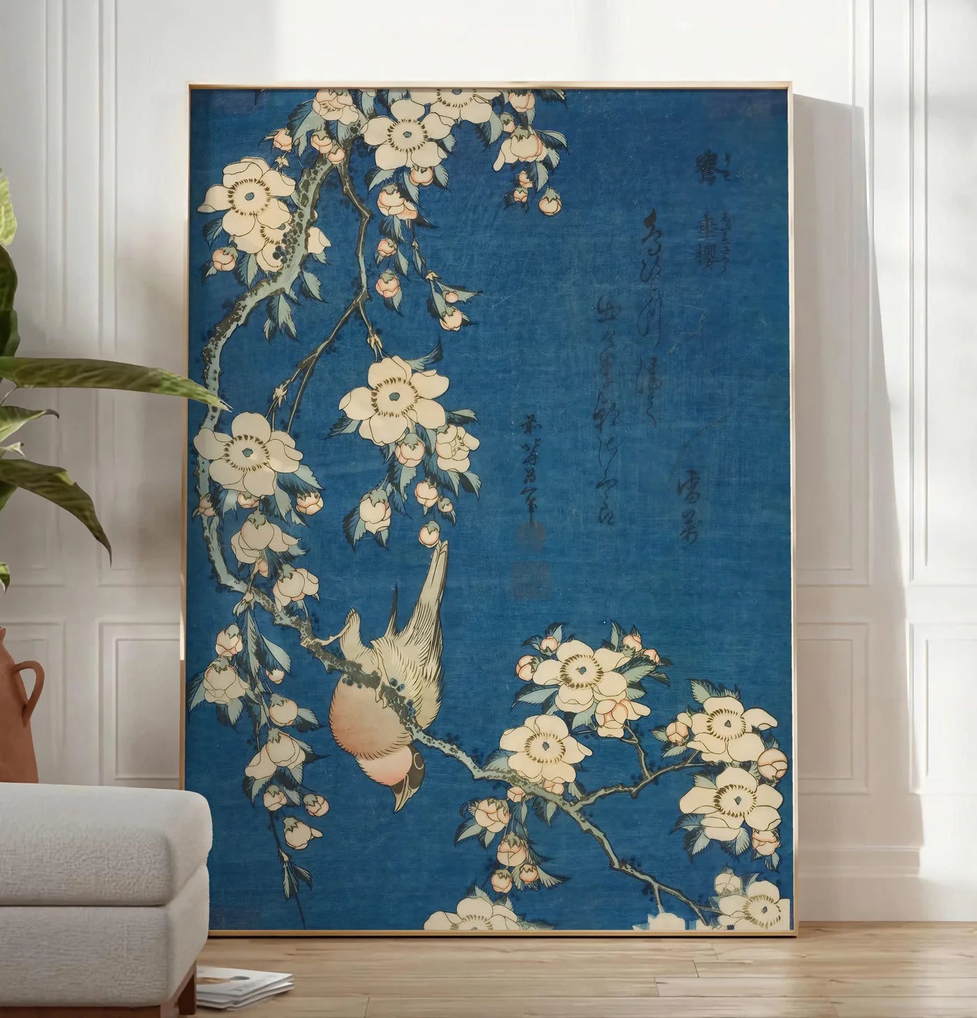 Reproduction affiche Hokusai bouvreuil sur cerisier, posee au sol dans un salon minimaliste clair, a cote d'un canape beige et d'une plante verte, sur un parquet clair