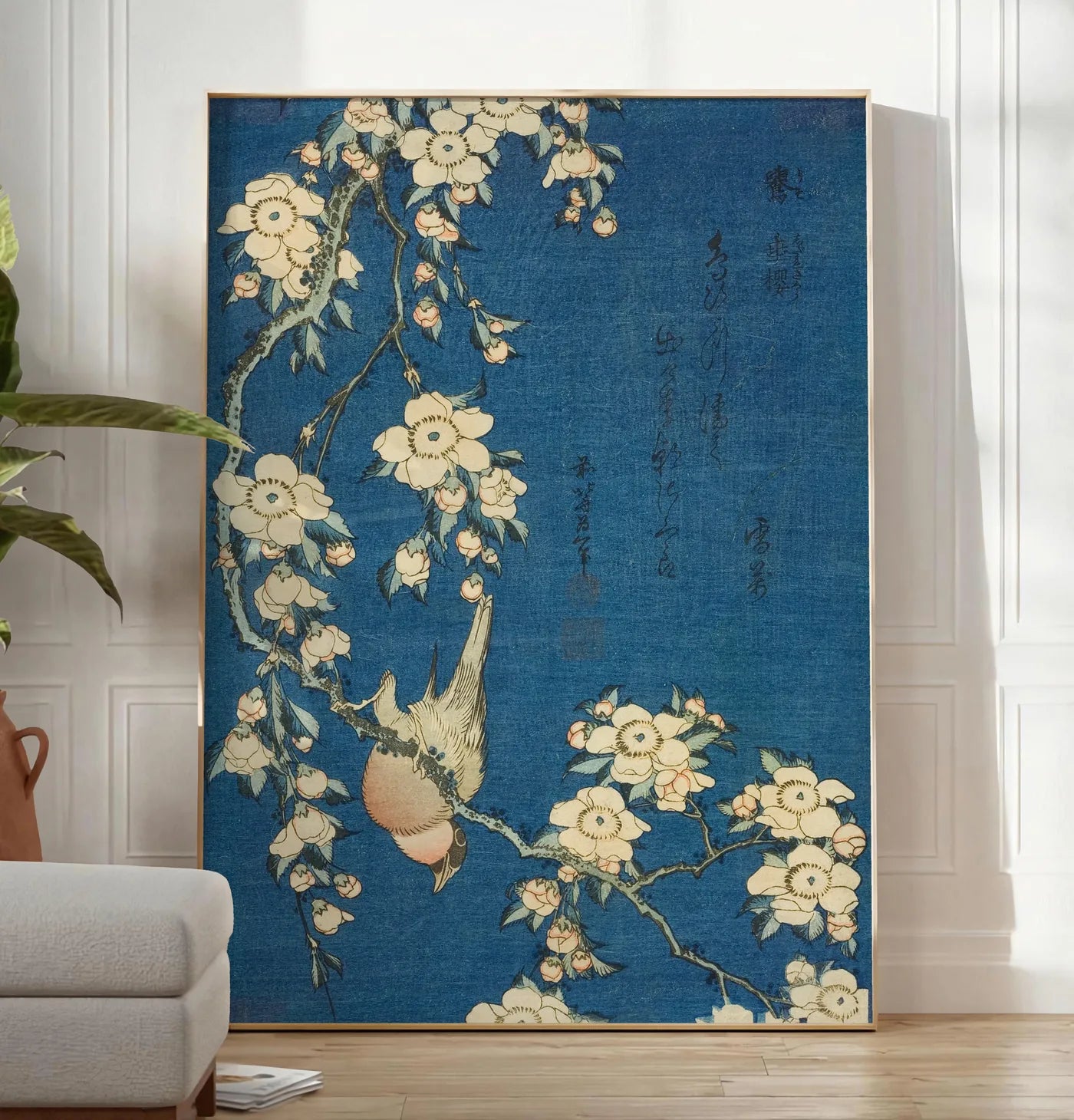 Reproduction affiche Hokusai cerisier en fleurs et bouvreuil, posee au sol dans un salon minimaliste clair, a cote d'un canape beige et d'une plante verte, sur un parquet clair