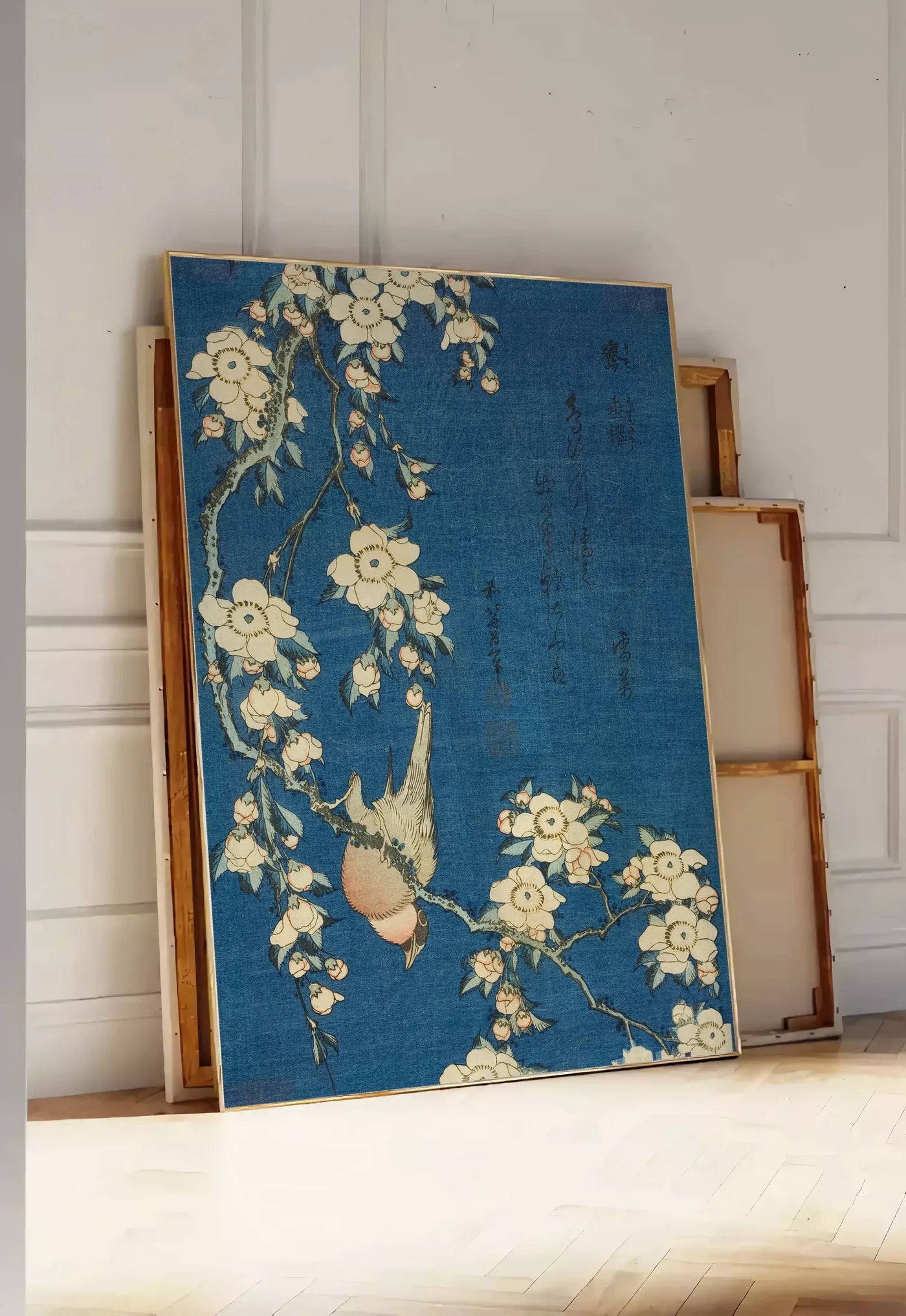 Tirage encadrable affiche Hokusai cerisier en fleurs et bouvreuil, posee au sol dans une entree lumineuse, contre un mur a panneaux blancs, a cote de chassis en bois empiles