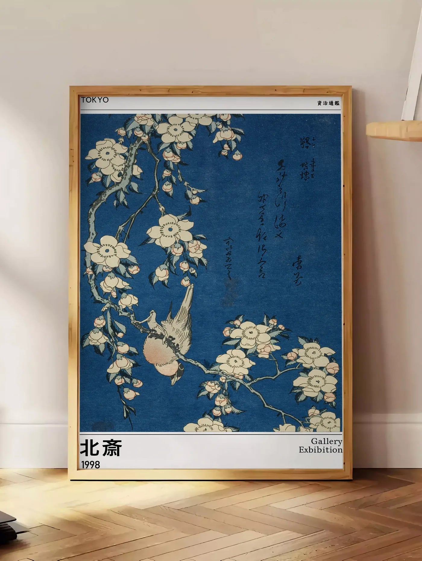 Estampe affiche Hokusai bouvreuil et cerisier, posee au sol dans un coin salon, sur un parquet a chevrons, pres d'une table d'appoint en chene chargee de livres