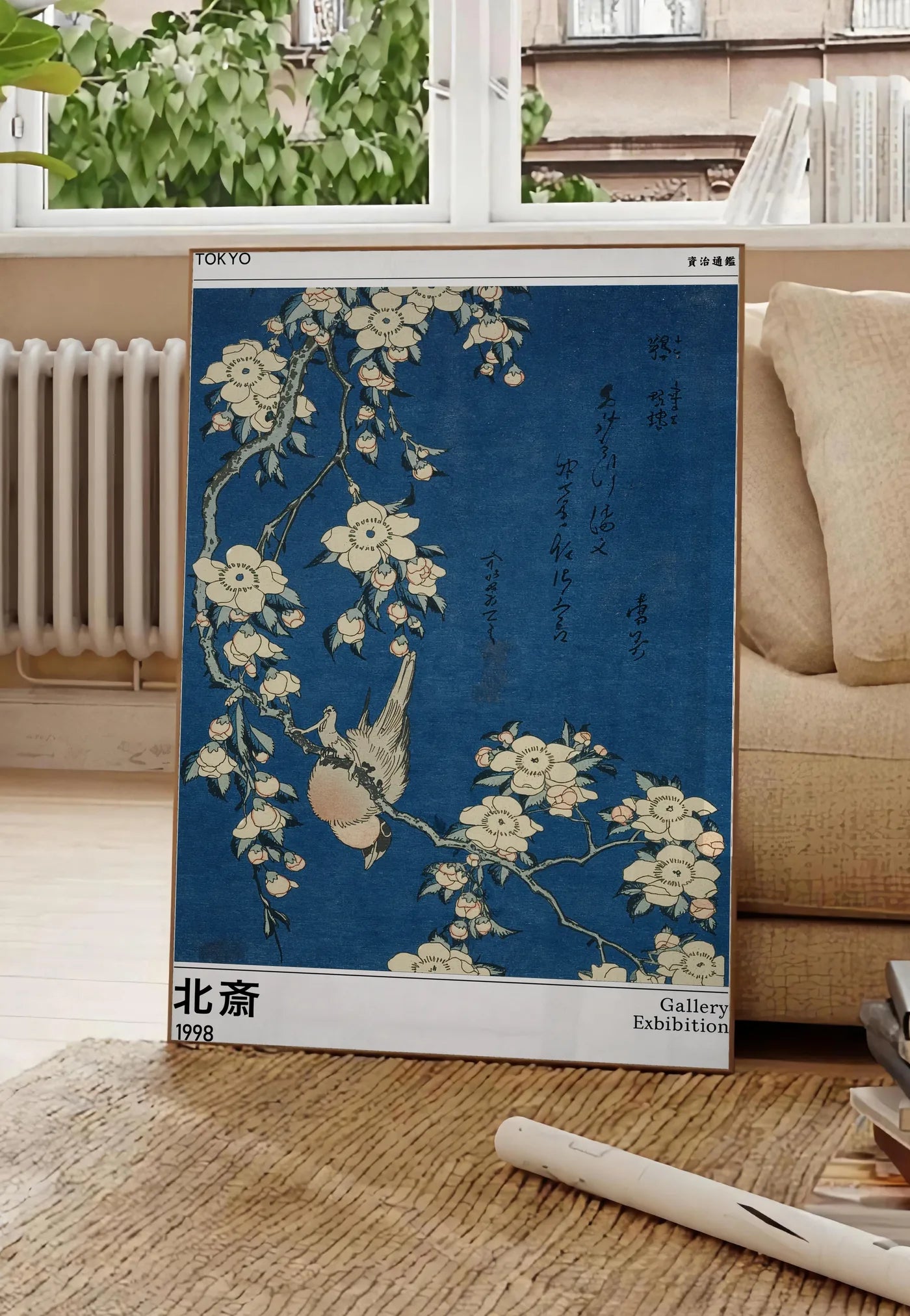 Poster affiche Hokusai bouvreuil et cerisier, posee au sol dans un salon parisien, devant une grande fenetre donnant sur un immeuble haussmannien, entre un ficus et un canape creme