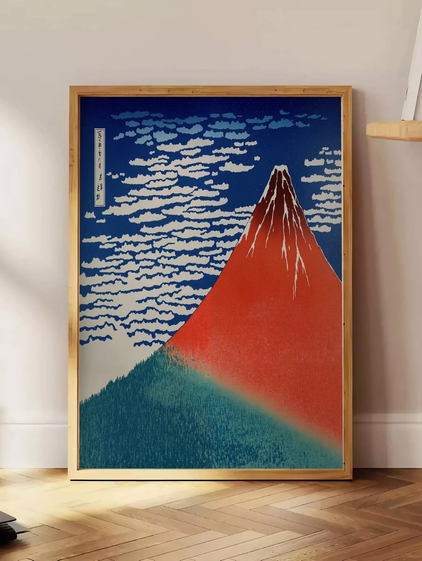 Estampe affiche mont Fuji rouge de Hokusai art japonais, posee au sol dans un coin salon, sur un parquet a chevrons, pres d'une table d'appoint en chene chargee de livres