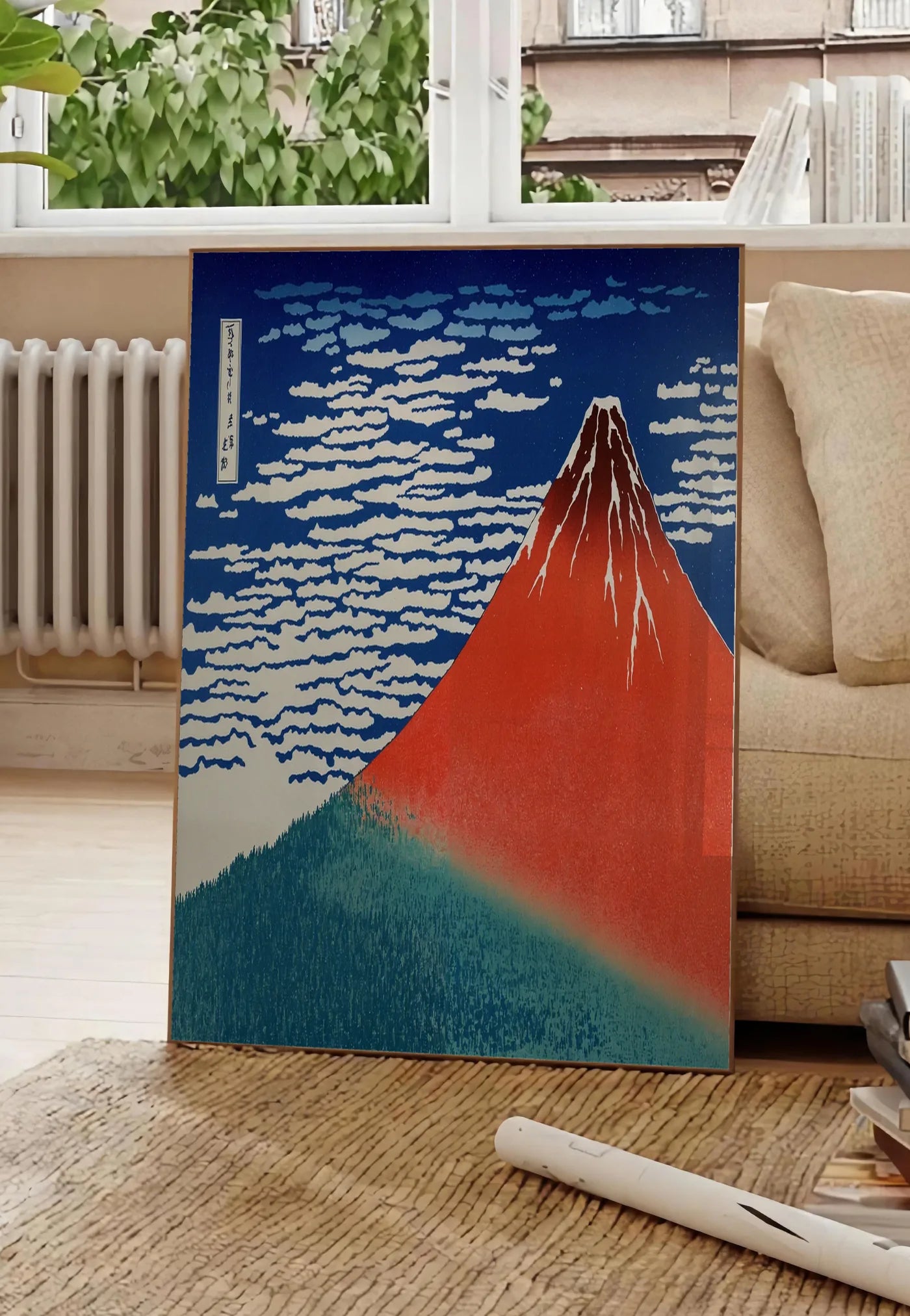 Poster affiche mont Fuji rouge de Hokusai art japonais, posee au sol dans un salon parisien, devant une grande fenetre donnant sur un immeuble haussmannien, entre un ficus et un canape creme