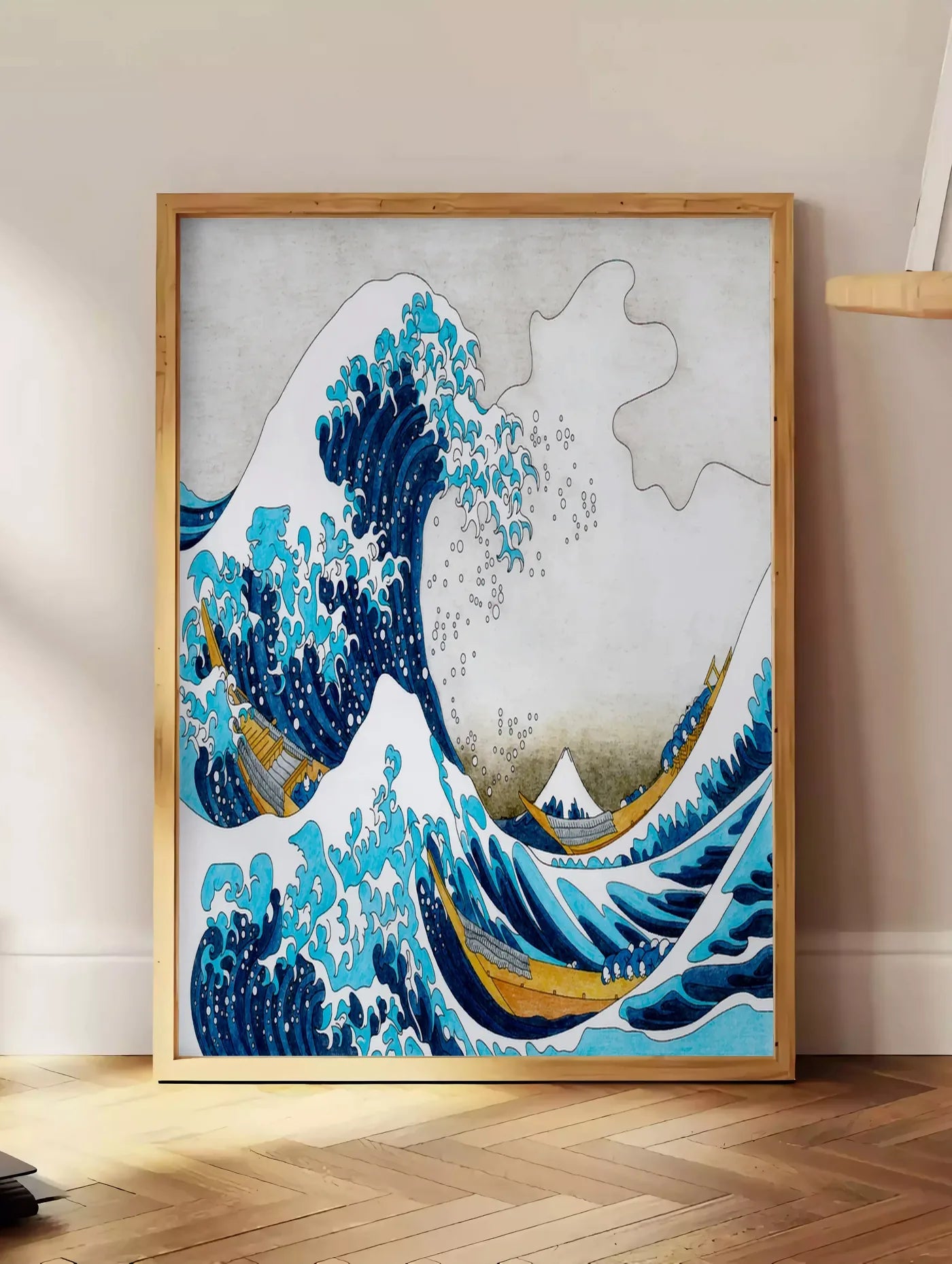 Estampe affiche Hokusai grande vague coloree, posee au sol dans un coin salon, sur un parquet a chevrons, pres d'une table d'appoint en chene chargee de livres