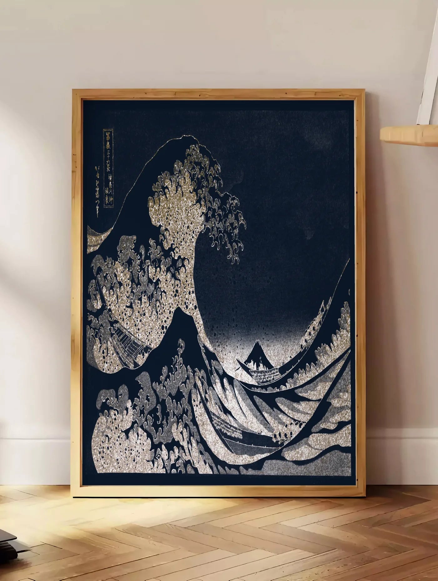 Estampe affiche Hokusai grande vague doree sur bleu nuit, posee au sol dans un coin salon, sur un parquet a chevrons, pres d'une table d'appoint en chene chargee de livres
