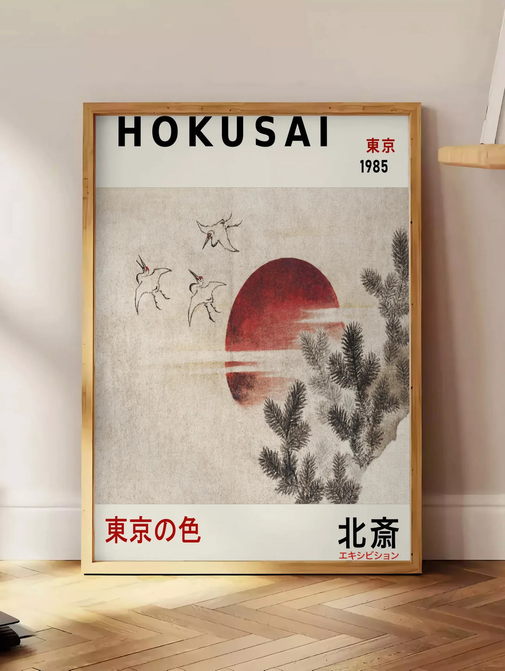 Estampe affiche Hokusai oiseaux en vol devant un soleil rouge, posee au sol dans un coin salon, sur un parquet a chevrons, pres d'une table d'appoint en chene chargee de livres