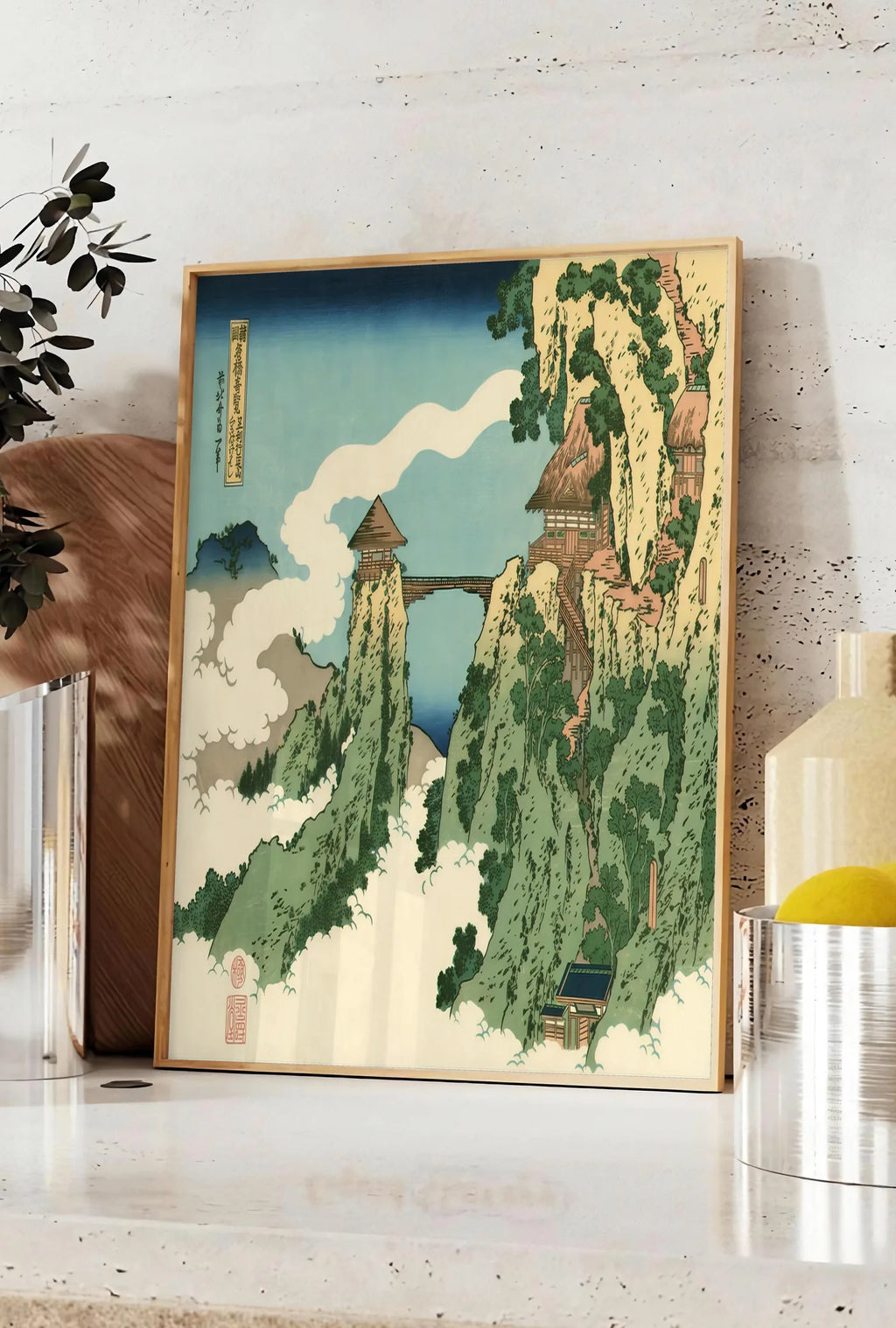 Affiche affiche Hokusai pont suspendu entre falaises au-dessus des nuages, posee sur un plan de travail de cuisine en pierre claire, entre une branche d'eucalyptus en vase et un bocal de citrons