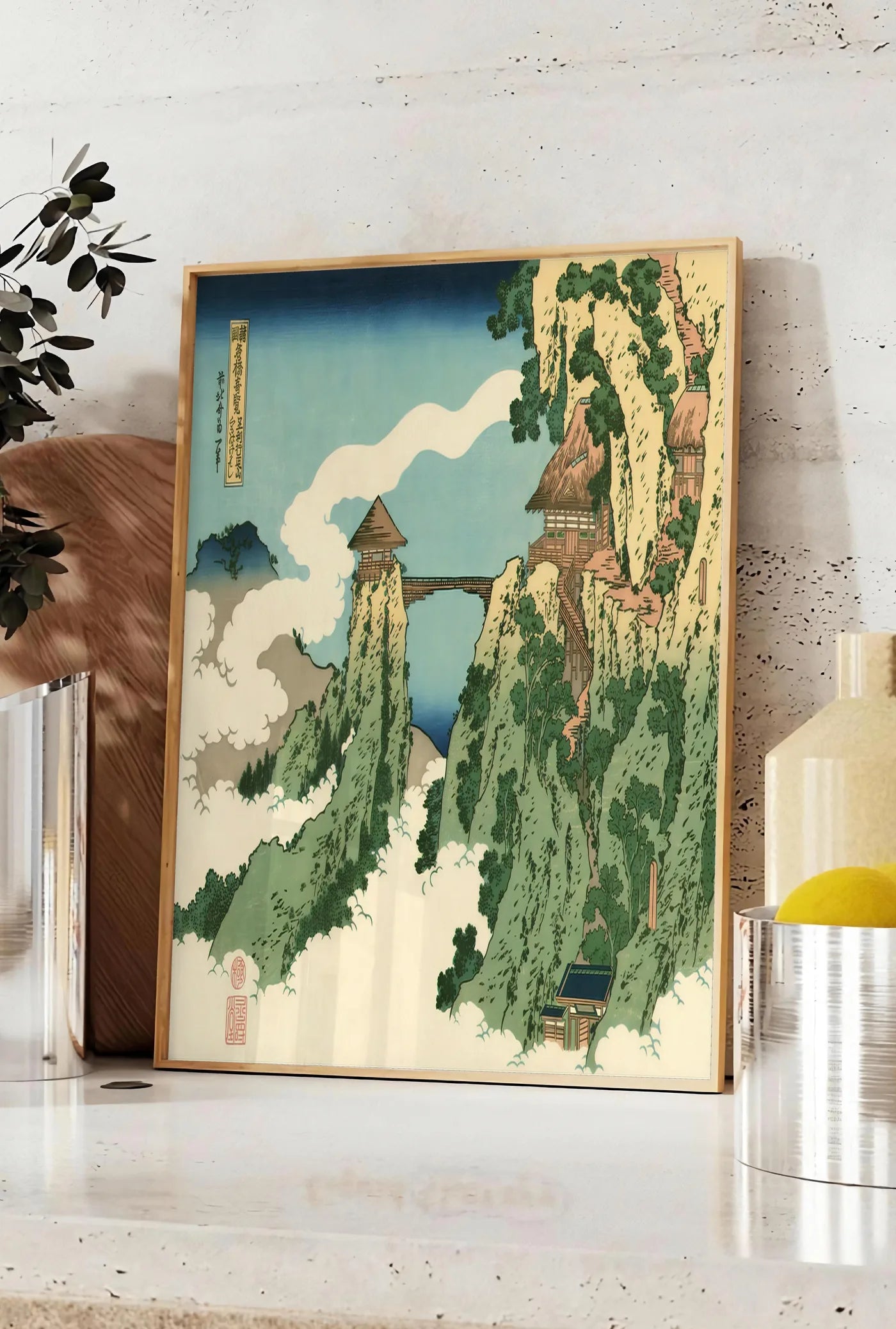 Affiche affiche Hokusai pont suspendu entre falaises au-dessus des nuages, posee sur un plan de travail de cuisine en pierre claire, entre une branche d'eucalyptus en vase et un bocal de citrons