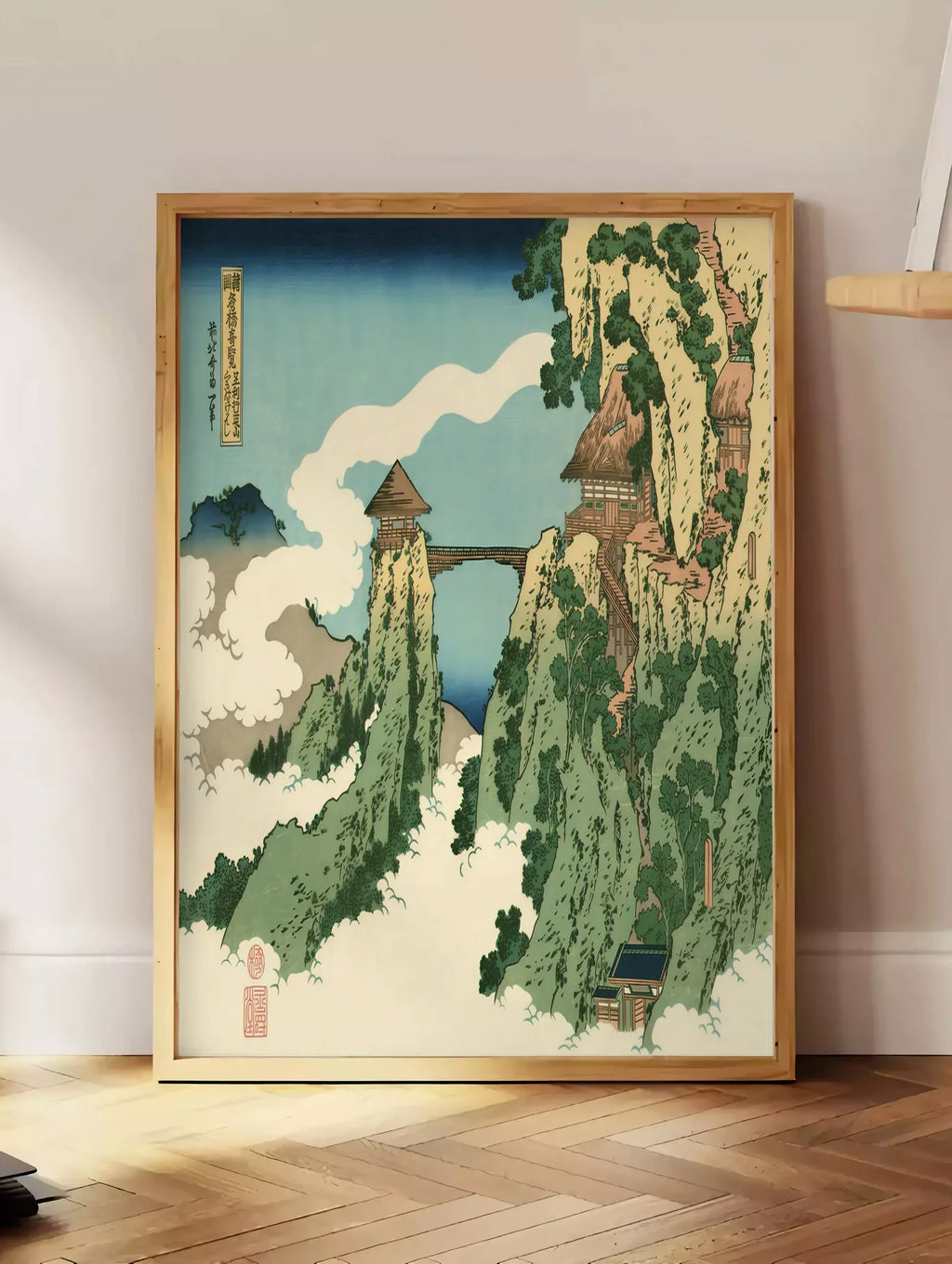 Estampe affiche Hokusai pont suspendu entre falaises au-dessus des nuages, posee au sol dans un coin salon, sur un parquet a chevrons, pres d'une table d'appoint en chene chargee de livres