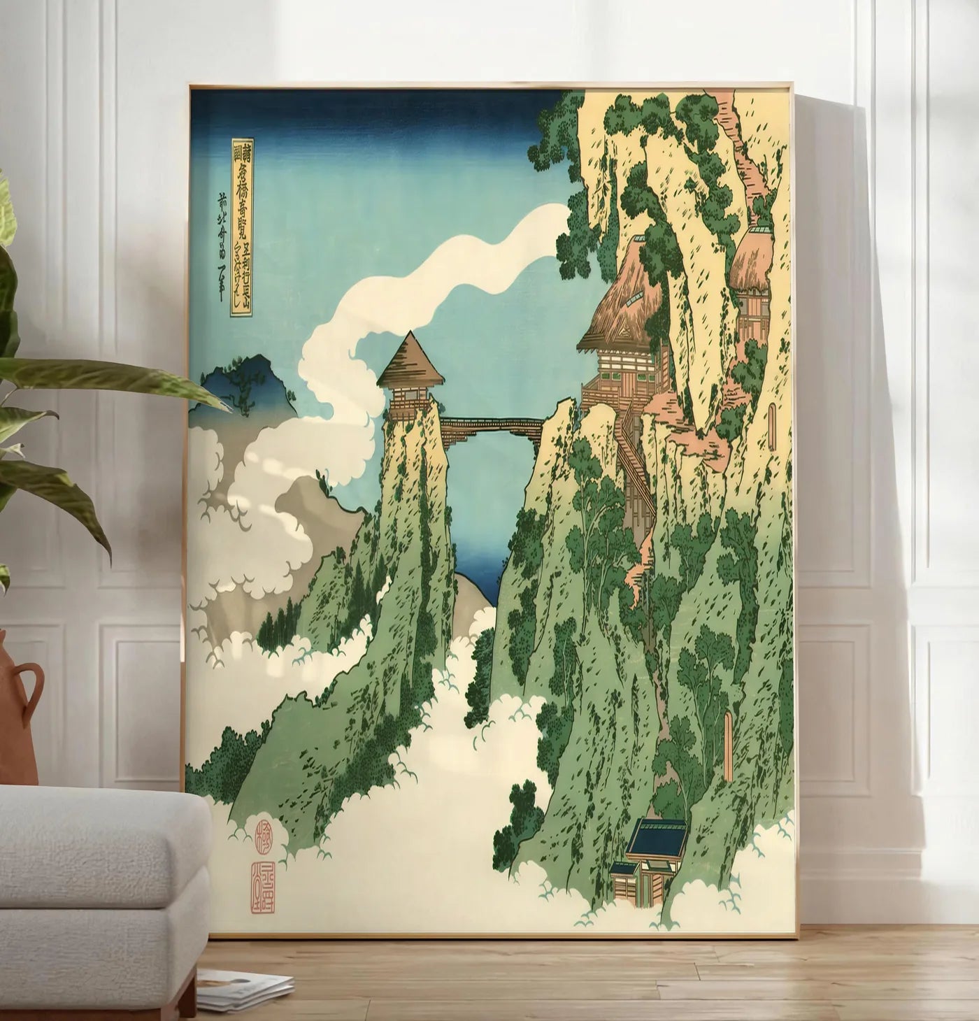 Reproduction affiche Hokusai pont suspendu entre falaises au-dessus des nuages, posee au sol dans un salon minimaliste clair, a cote d'un canape beige et d'une plante verte, sur un parquet clair