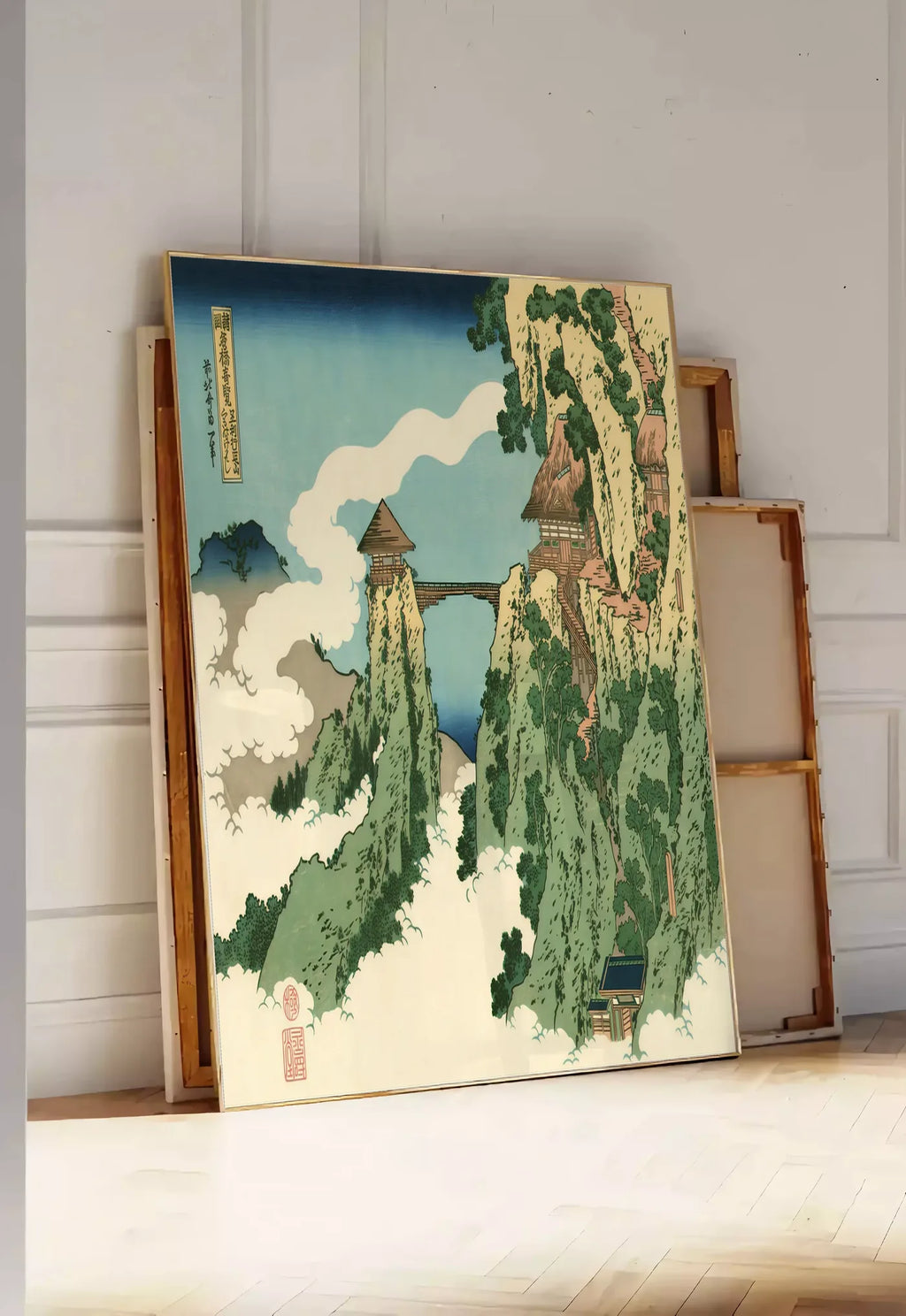 Tirage encadrable affiche Hokusai pont suspendu entre falaises au-dessus des nuages, posee au sol dans une entree lumineuse, contre un mur a panneaux blancs, a cote de chassis en bois empiles