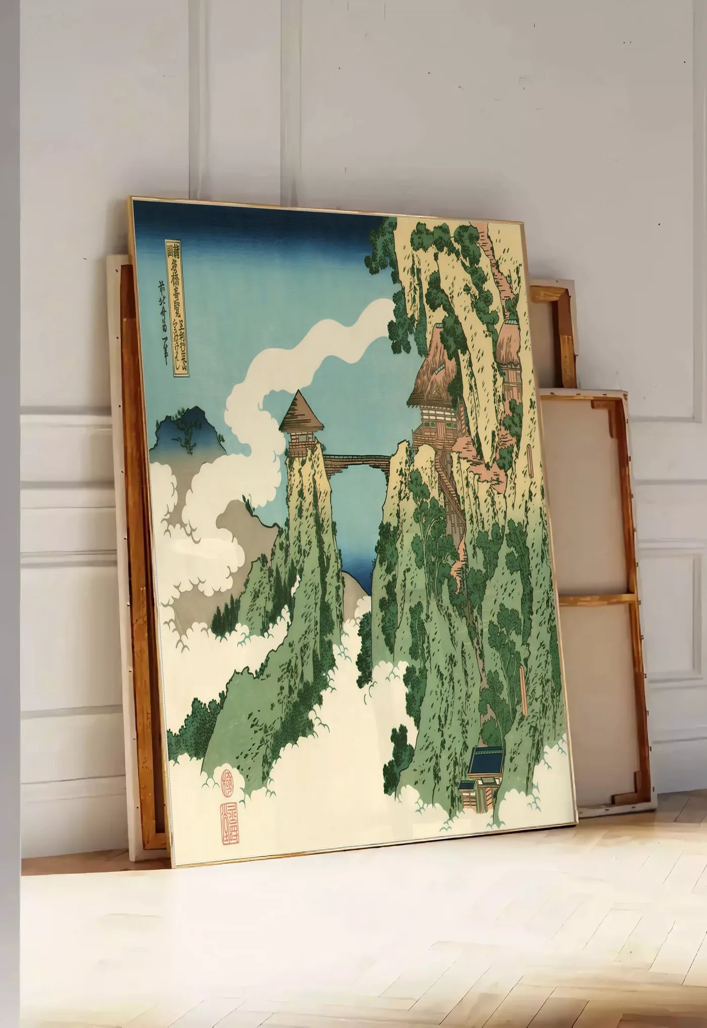Tirage encadrable affiche Hokusai pont suspendu entre falaises au-dessus des nuages, posee au sol dans une entree lumineuse, contre un mur a panneaux blancs, a cote de chassis en bois empiles