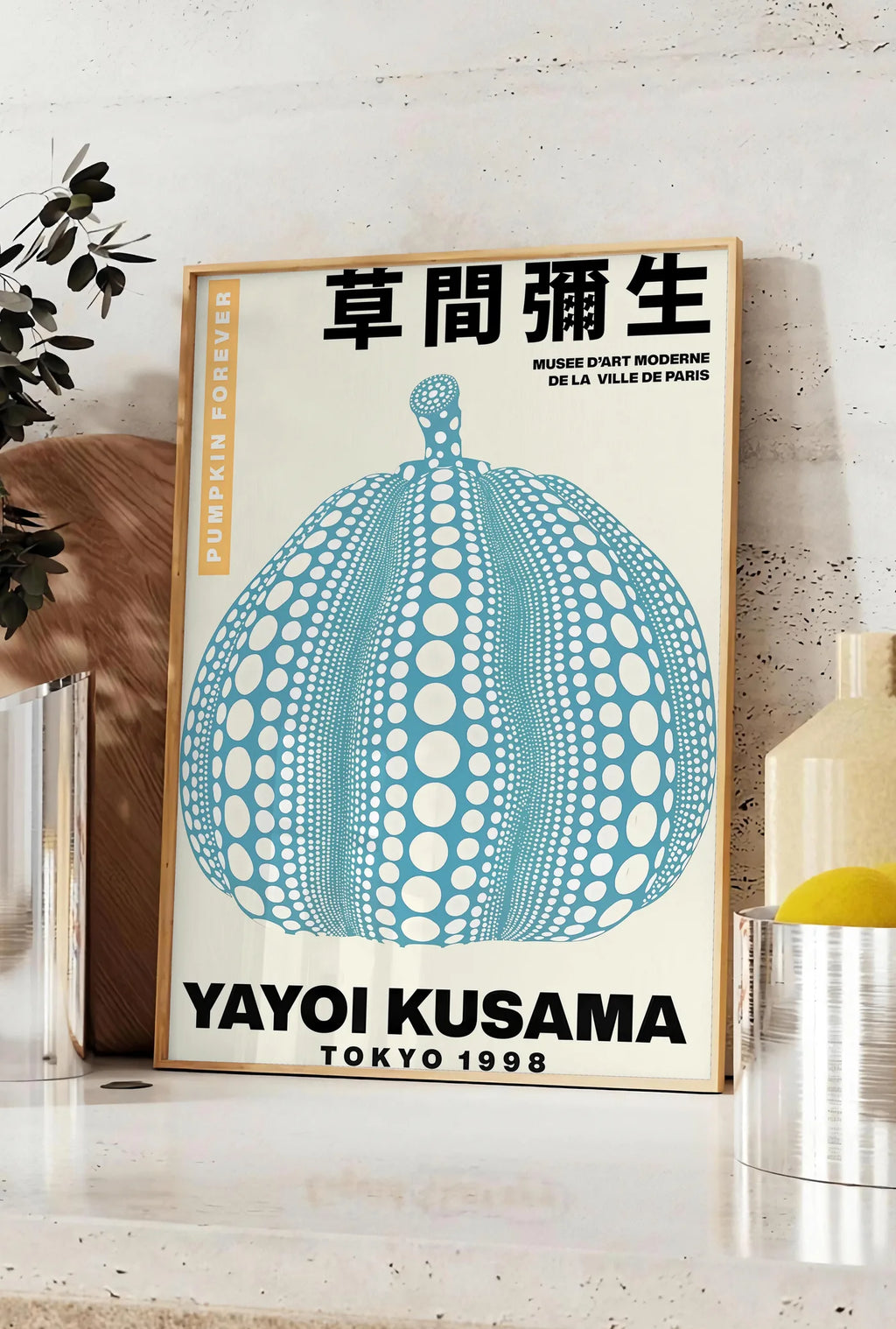 Affiche affiche Kusama citrouille bleue a pois sur fond creme, posee sur un plan de travail de cuisine en pierre claire, entre une branche d'eucalyptus en vase et un bocal de citrons