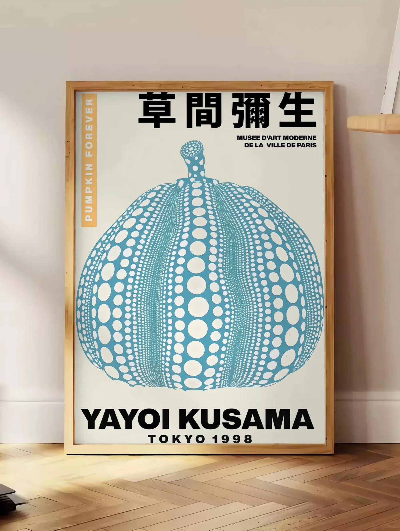 Estampe affiche Kusama citrouille bleue a pois sur fond creme, posee au sol dans un coin salon, sur un parquet a chevrons, pres d'une table d'appoint en chene chargee de livres