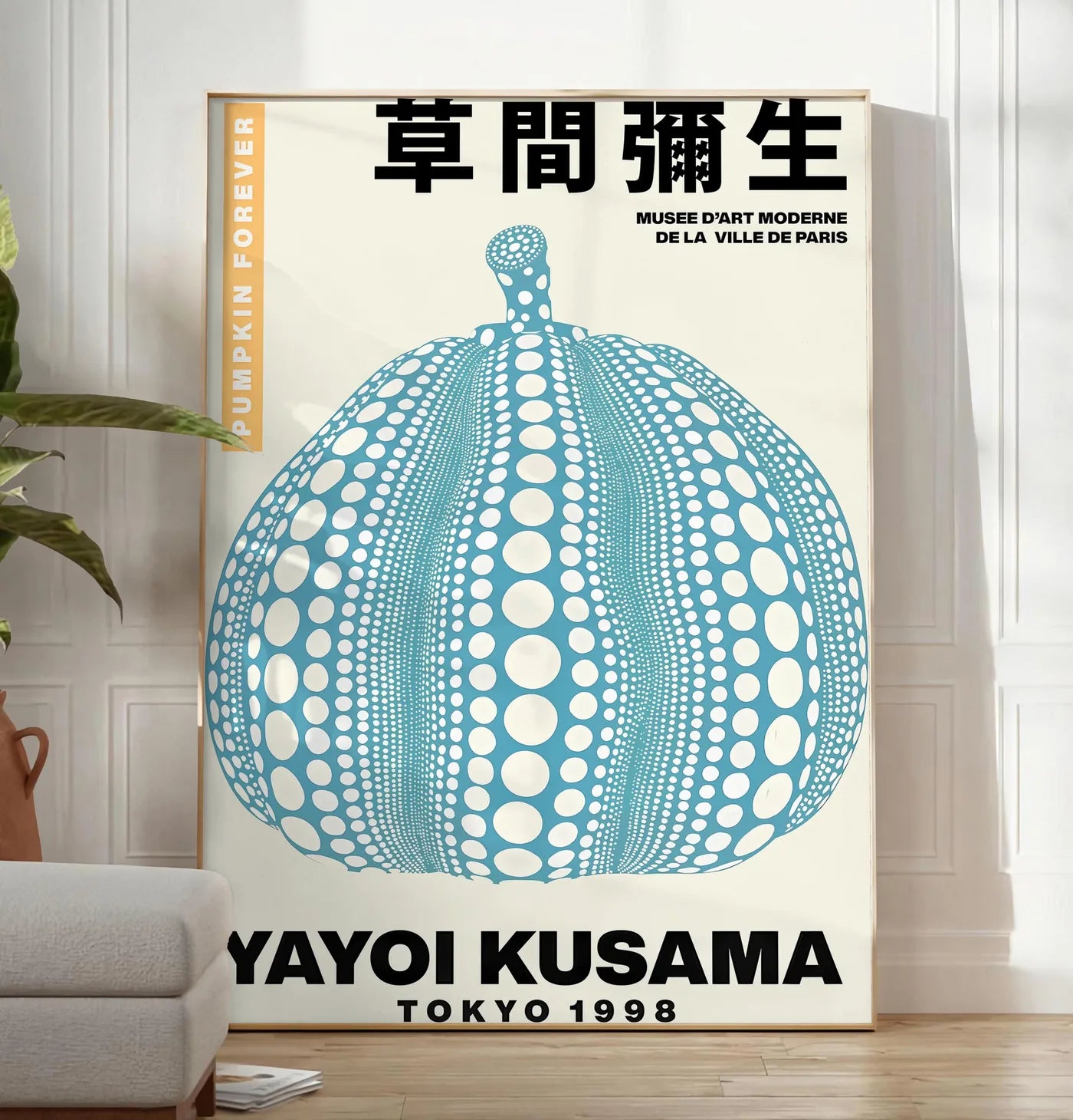Reproduction affiche Kusama citrouille bleue a pois sur fond creme, posee au sol dans un salon minimaliste clair, a cote d'un canape beige et d'une plante verte, sur un parquet clair