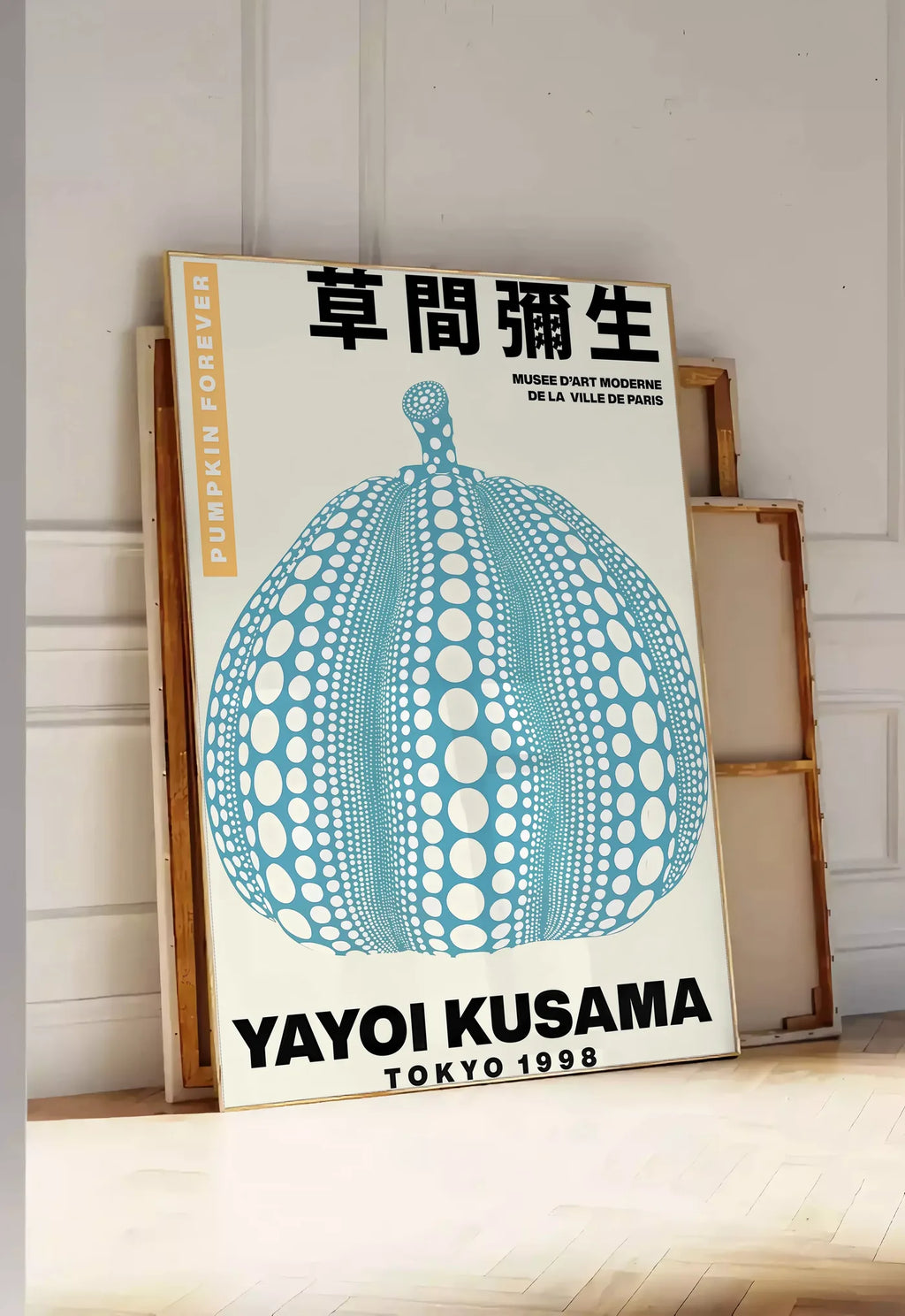 Tirage encadrable affiche Kusama citrouille bleue a pois sur fond creme, posee au sol dans une entree lumineuse, contre un mur a panneaux blancs, a cote de chassis en bois empiles