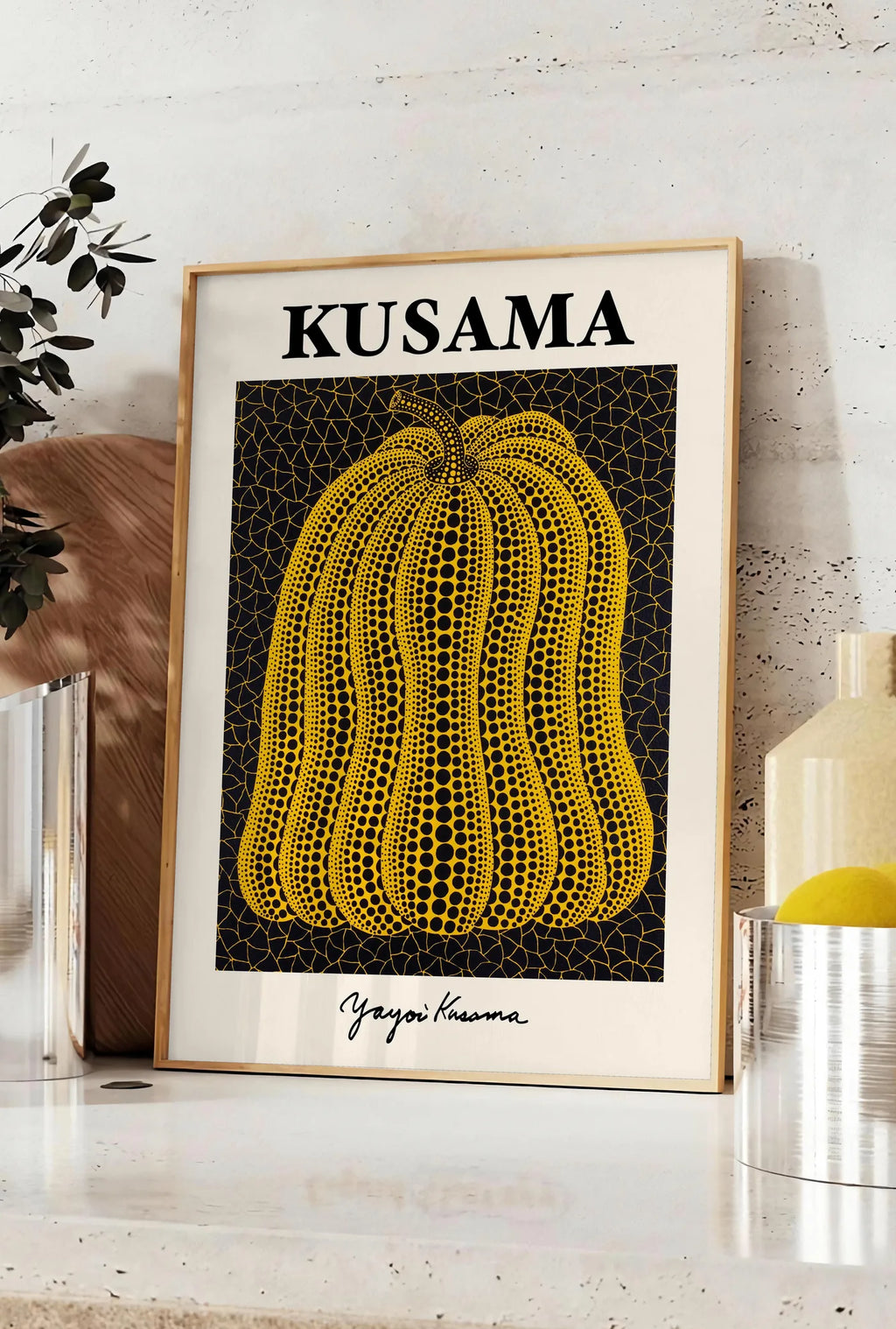 Affiche affiche Kusama citrouille jaune a pois sur fond noir signe, posee sur un plan de travail de cuisine en pierre claire, entre une branche d'eucalyptus en vase et un bocal de citrons