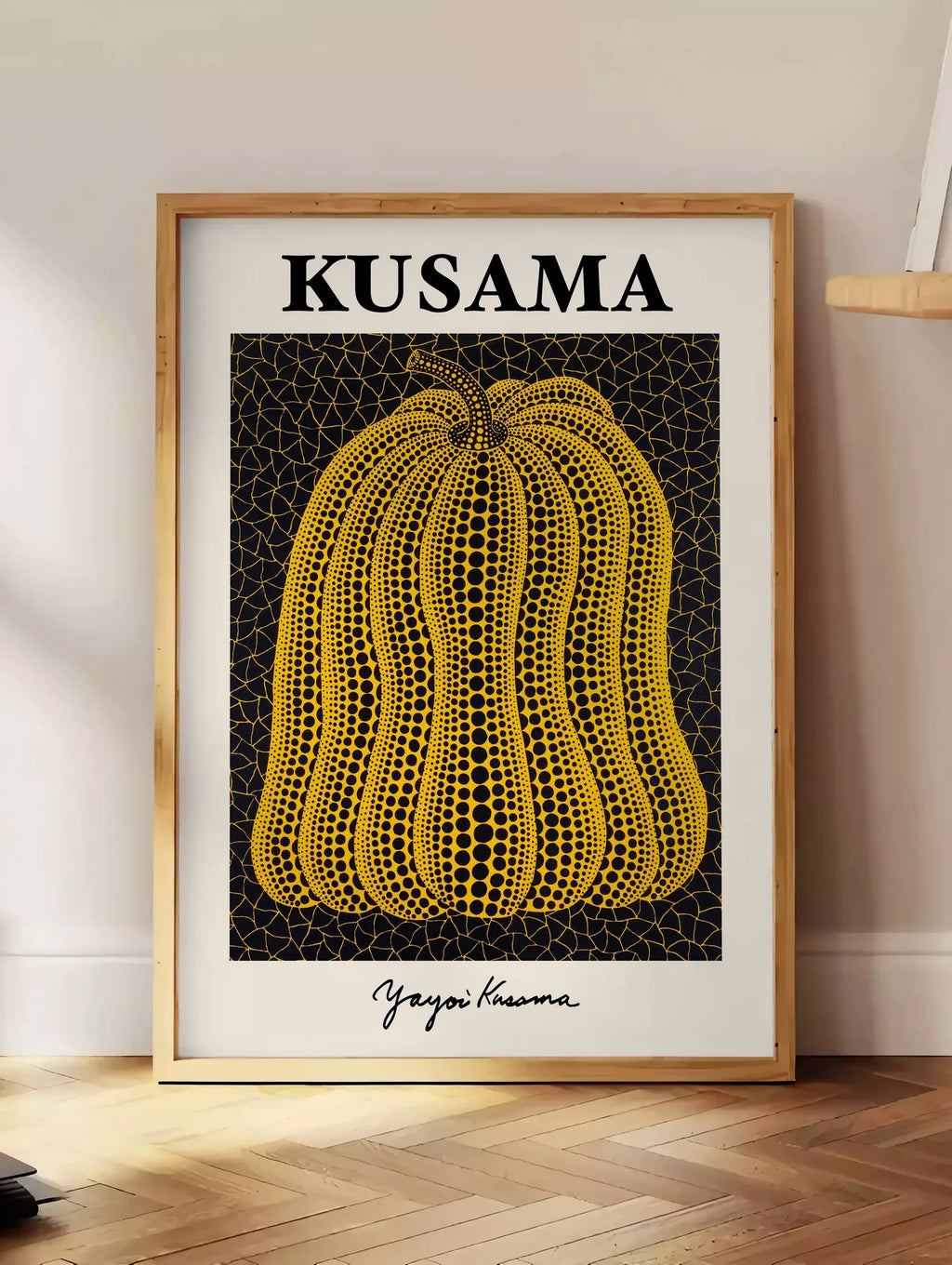 Estampe affiche Kusama citrouille jaune a pois sur fond noir signe, posee au sol dans un coin salon, sur un parquet a chevrons, pres d'une table d'appoint en chene chargee de livres