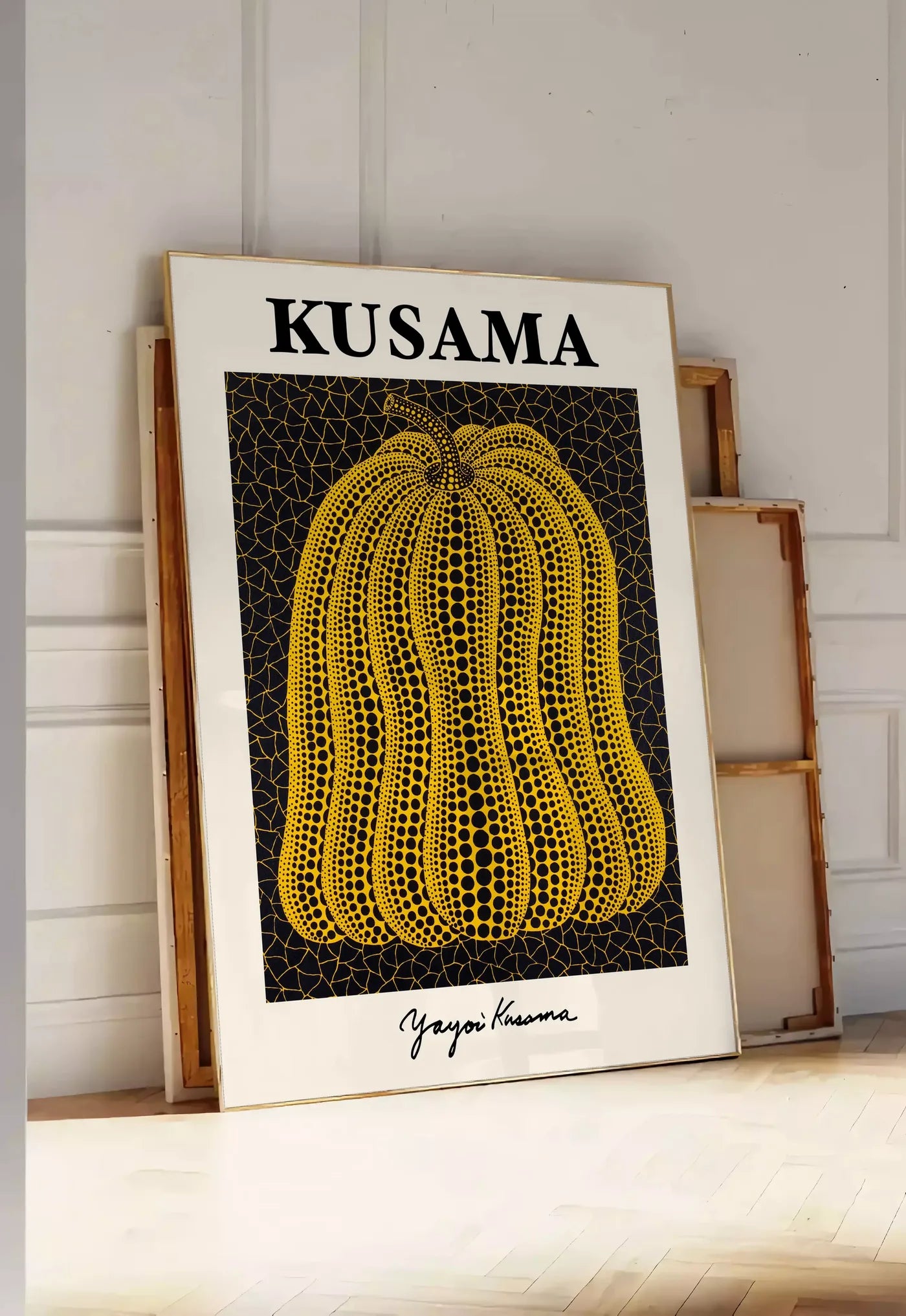 Tirage encadrable affiche Kusama citrouille jaune a pois sur fond noir signe, posee au sol dans une entree lumineuse, contre un mur a panneaux blancs, a cote de chassis en bois empiles