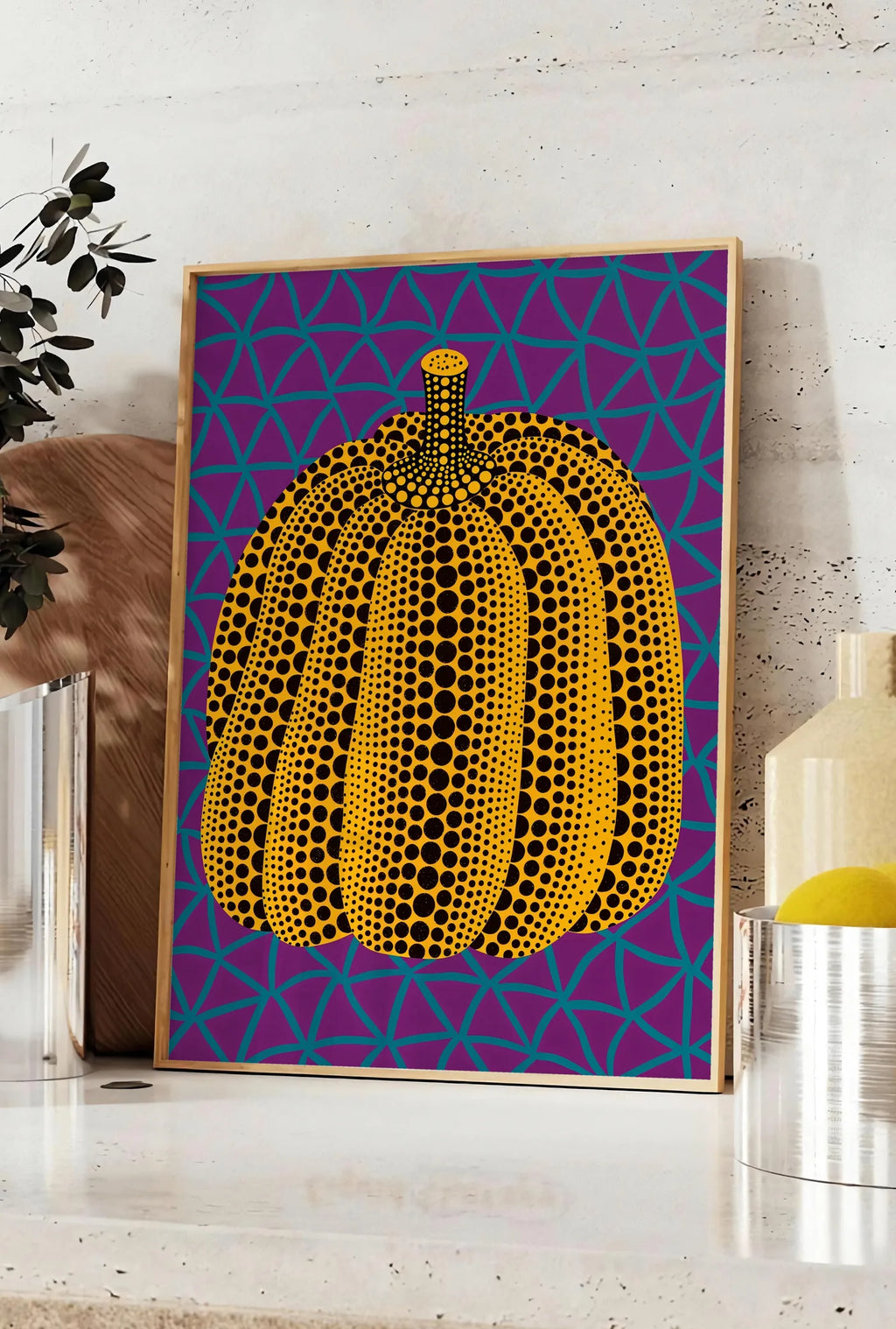 Affiche affiche Kusama citrouille jaune a pois sur fond violet a filet bleu, posee sur un plan de travail de cuisine en pierre claire, entre une branche d'eucalyptus en vase et un bocal de citrons