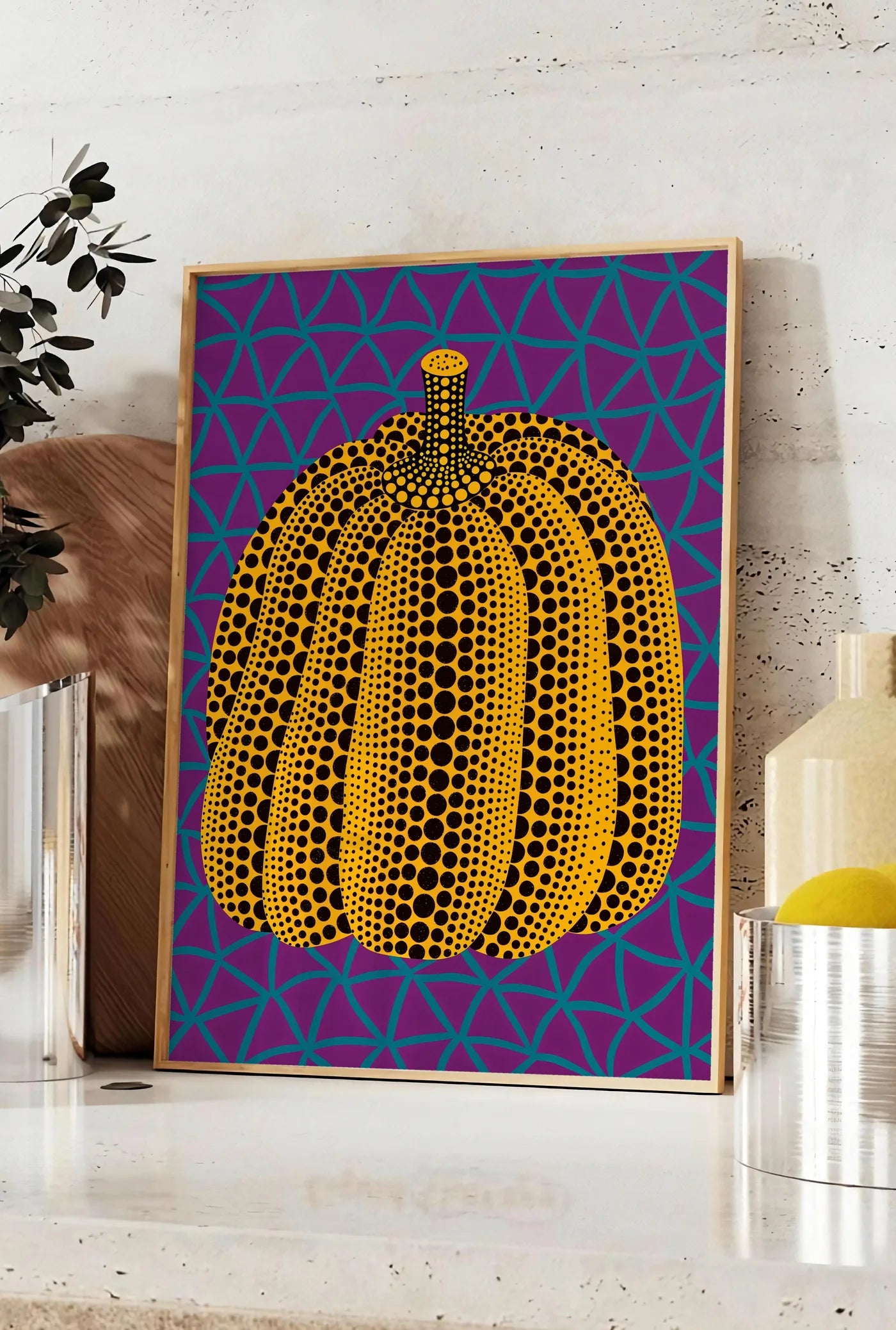 Affiche affiche Kusama citrouille jaune a pois sur fond violet a filet bleu, posee sur un plan de travail de cuisine en pierre claire, entre une branche d'eucalyptus en vase et un bocal de citrons