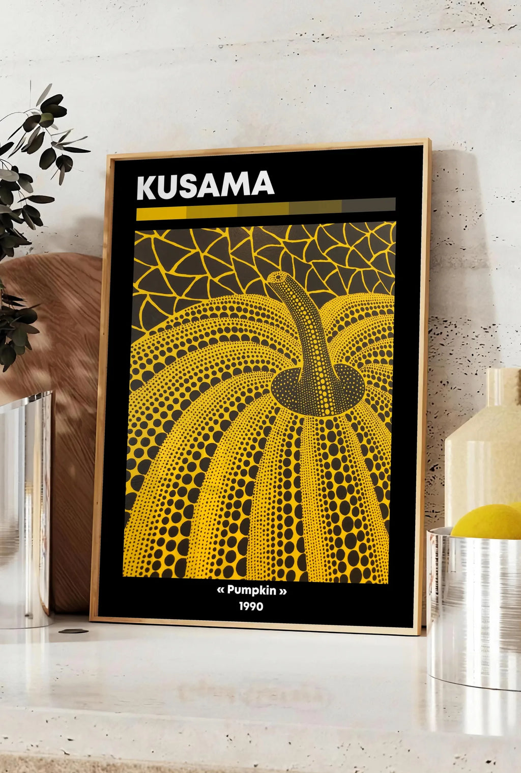 Affiche affiche Kusama citrouille jaune a pois vue en contre-plongee sur fond noir, posee sur un plan de travail de cuisine en pierre claire, entre une branche d'eucalyptus en vase et un bocal de citrons