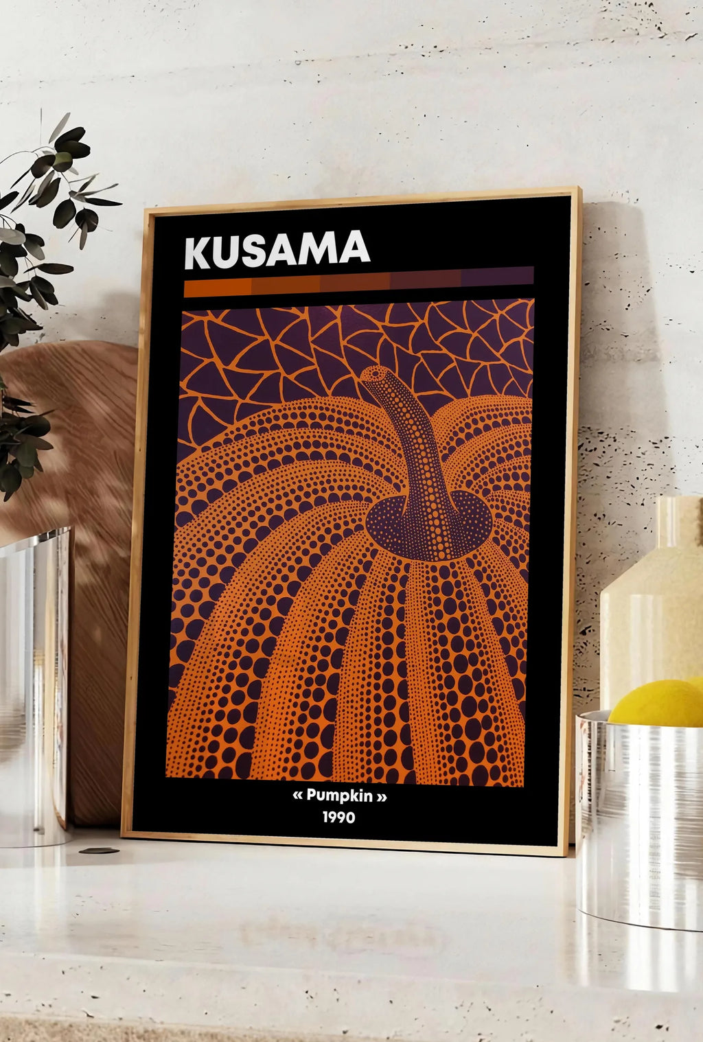 Affiche affiche Kusama citrouille orange a pois vue en contre-plongee sur fond violet, posee sur un plan de travail de cuisine en pierre claire, entre une branche d'eucalyptus en vase et un bocal de citrons