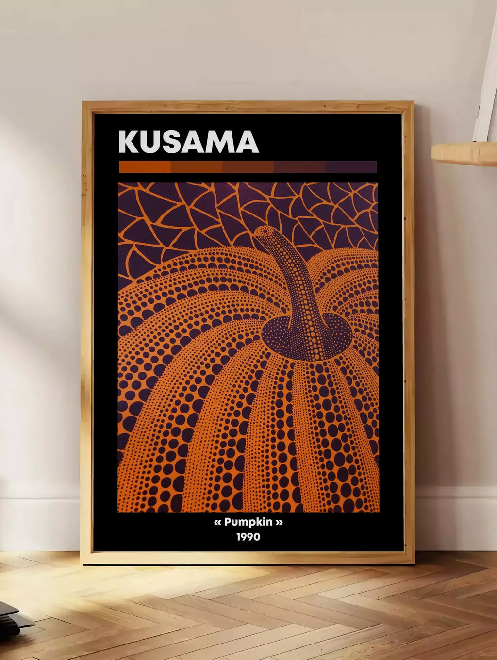 Estampe affiche Kusama citrouille orange a pois vue en contre-plongee sur fond violet, posee au sol dans un coin salon, sur un parquet a chevrons, pres d'une table d'appoint en chene chargee de livres