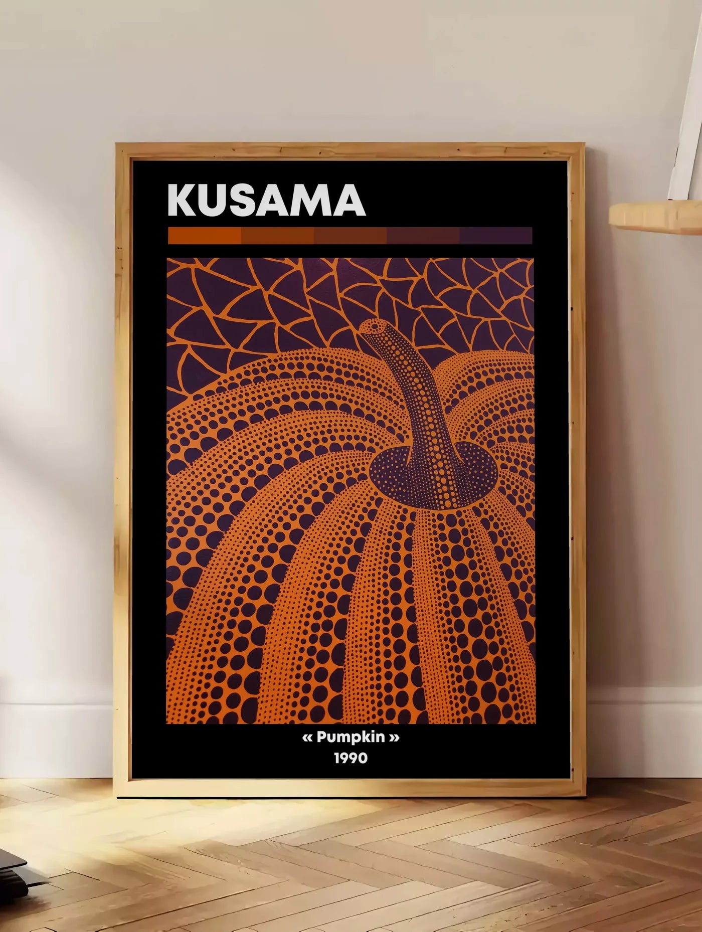 Estampe affiche Kusama citrouille orange a pois vue en contre-plongee sur fond violet, posee au sol dans un coin salon, sur un parquet a chevrons, pres d'une table d'appoint en chene chargee de livres