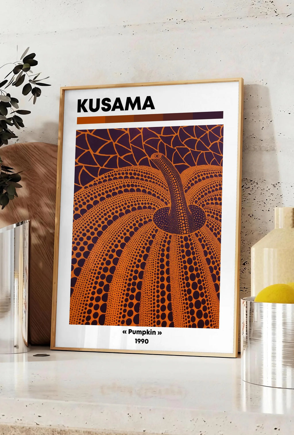 Affiche affiche Kusama citrouille orange feu a pois sur fond aubergine, posee sur un plan de travail de cuisine en pierre claire, entre une branche d'eucalyptus en vase et un bocal de citrons