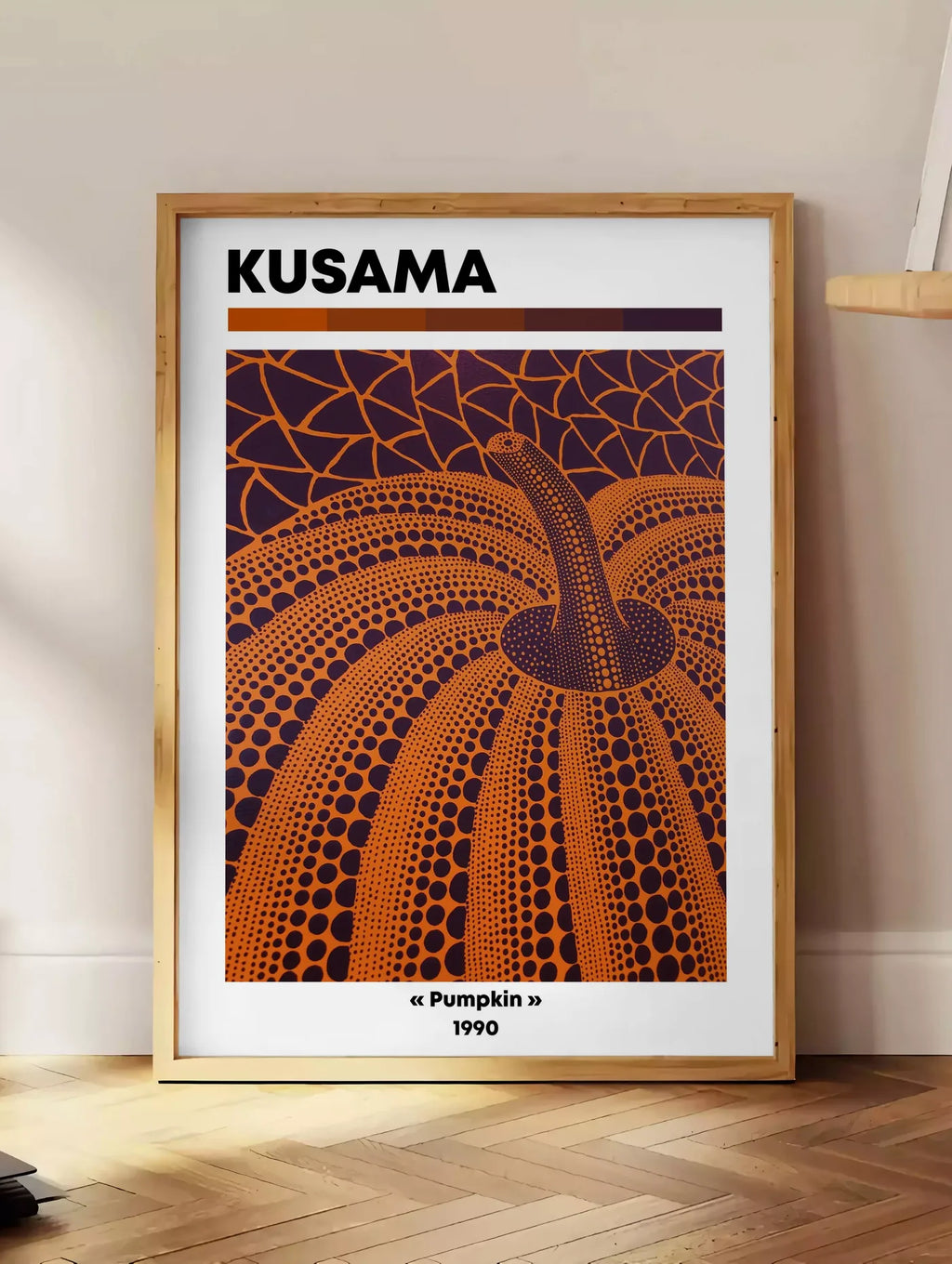 Estampe affiche Kusama citrouille orange feu a pois sur fond aubergine, posee au sol dans un coin salon, sur un parquet a chevrons, pres d'une table d'appoint en chene chargee de livres