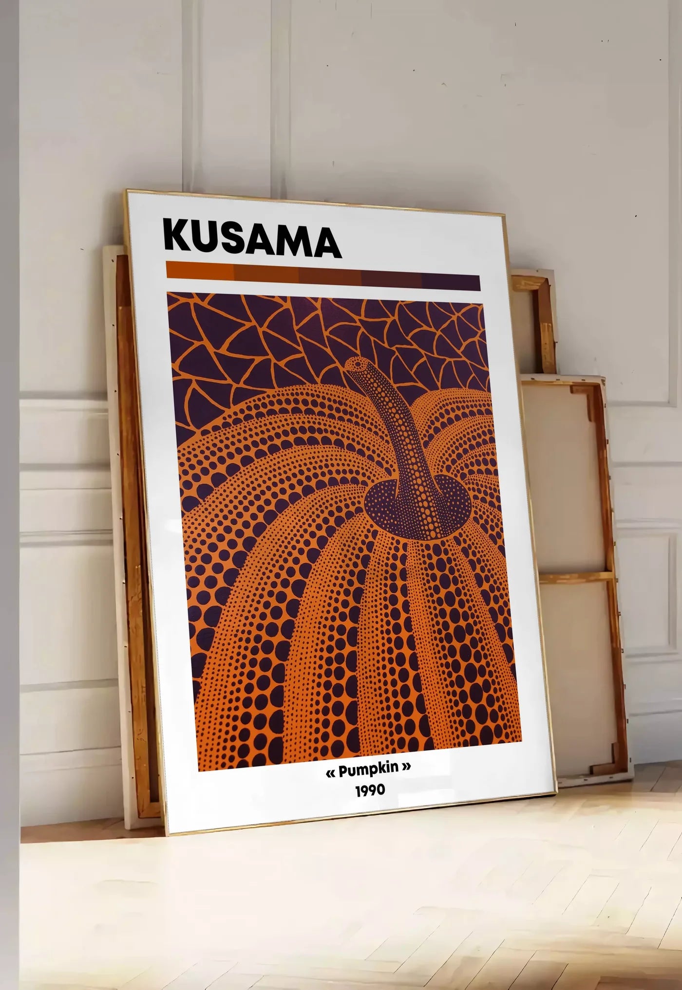 Tirage encadrable affiche Kusama citrouille orange feu a pois sur fond aubergine, posee au sol dans une entree lumineuse, contre un mur a panneaux blancs, a cote de chassis en bois empiles