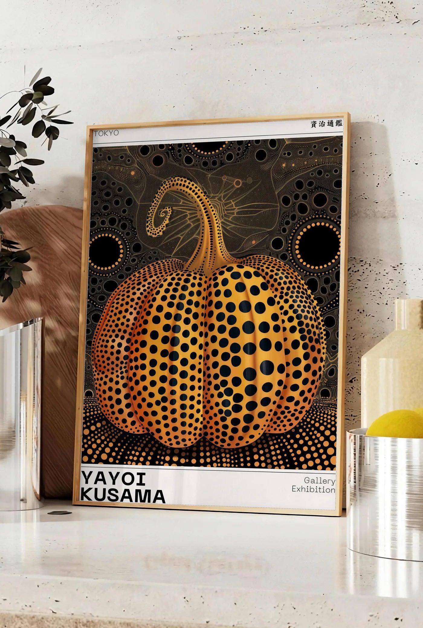 Affiche affiche Kusama citrouille orange a pois sur fond noir, posee sur un plan de travail de cuisine en pierre claire, entre une branche d'eucalyptus en vase et un bocal de citrons