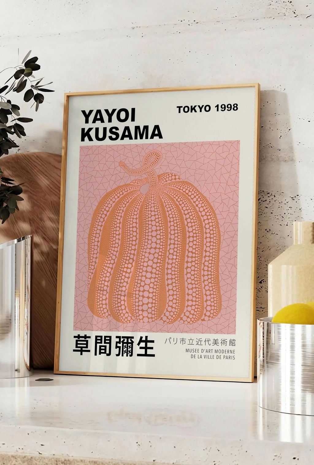 Affiche affiche Kusama citrouille orange a pois sur fond rose, posee sur un plan de travail de cuisine en pierre claire, entre une branche d'eucalyptus en vase et un bocal de citrons