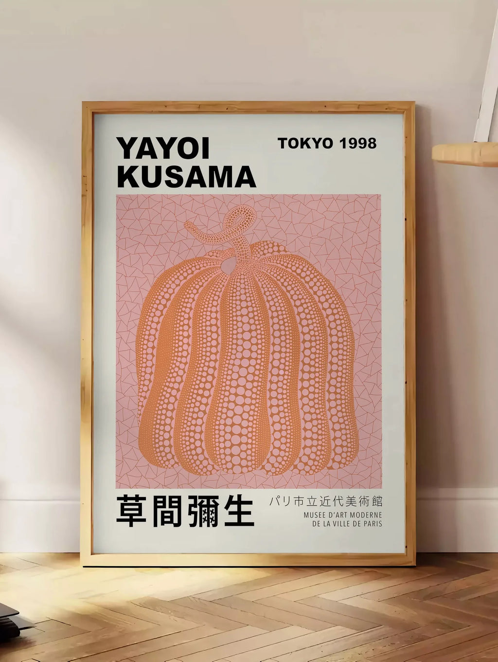 Estampe affiche Kusama citrouille orange a pois sur fond rose, posee au sol dans un coin salon, sur un parquet a chevrons, pres d'une table d'appoint en chene chargee de livres