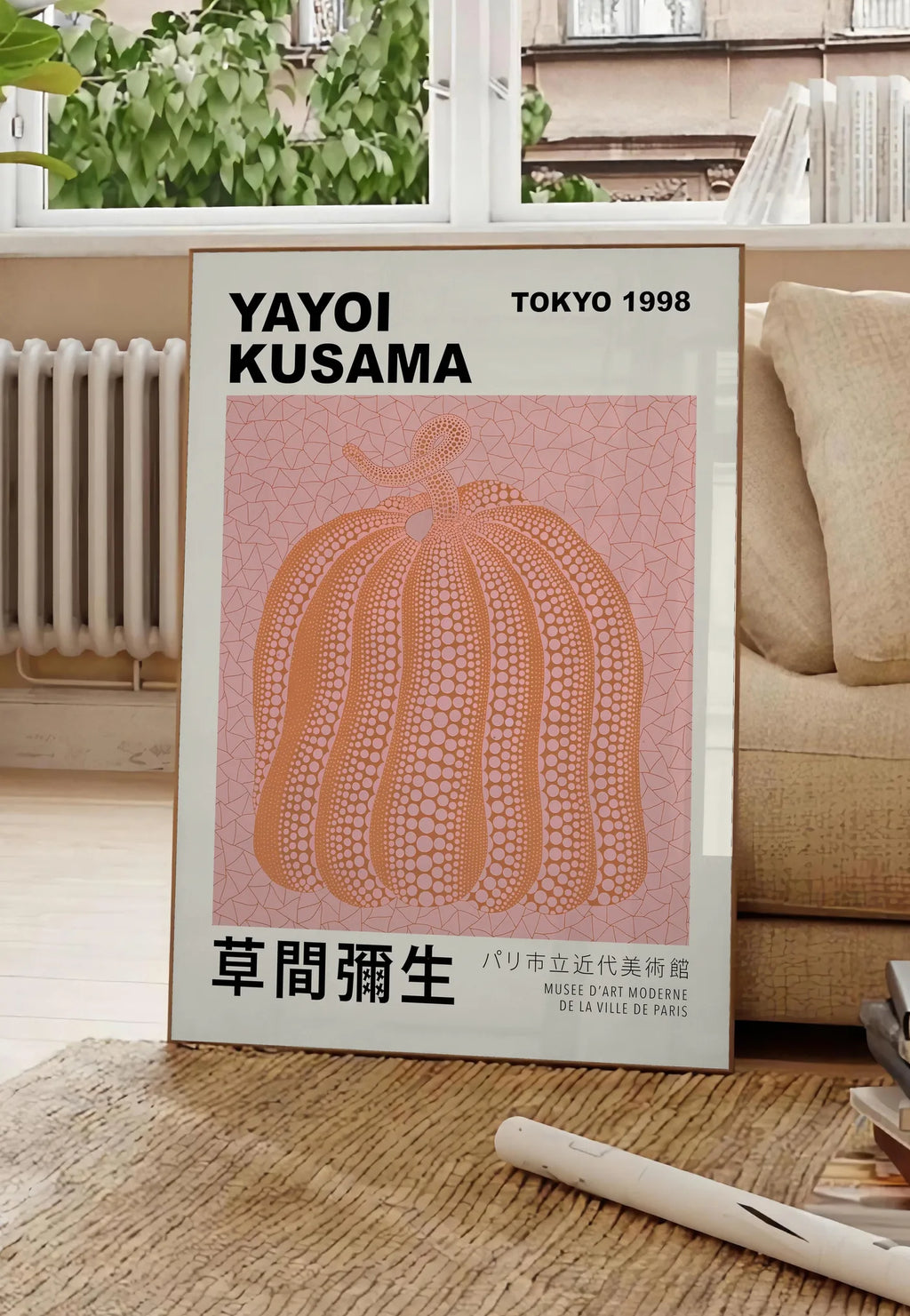 Poster affiche Kusama citrouille orange a pois sur fond rose, posee au sol dans un salon parisien, devant une grande fenetre donnant sur un immeuble haussmannien, entre un ficus et un canape creme