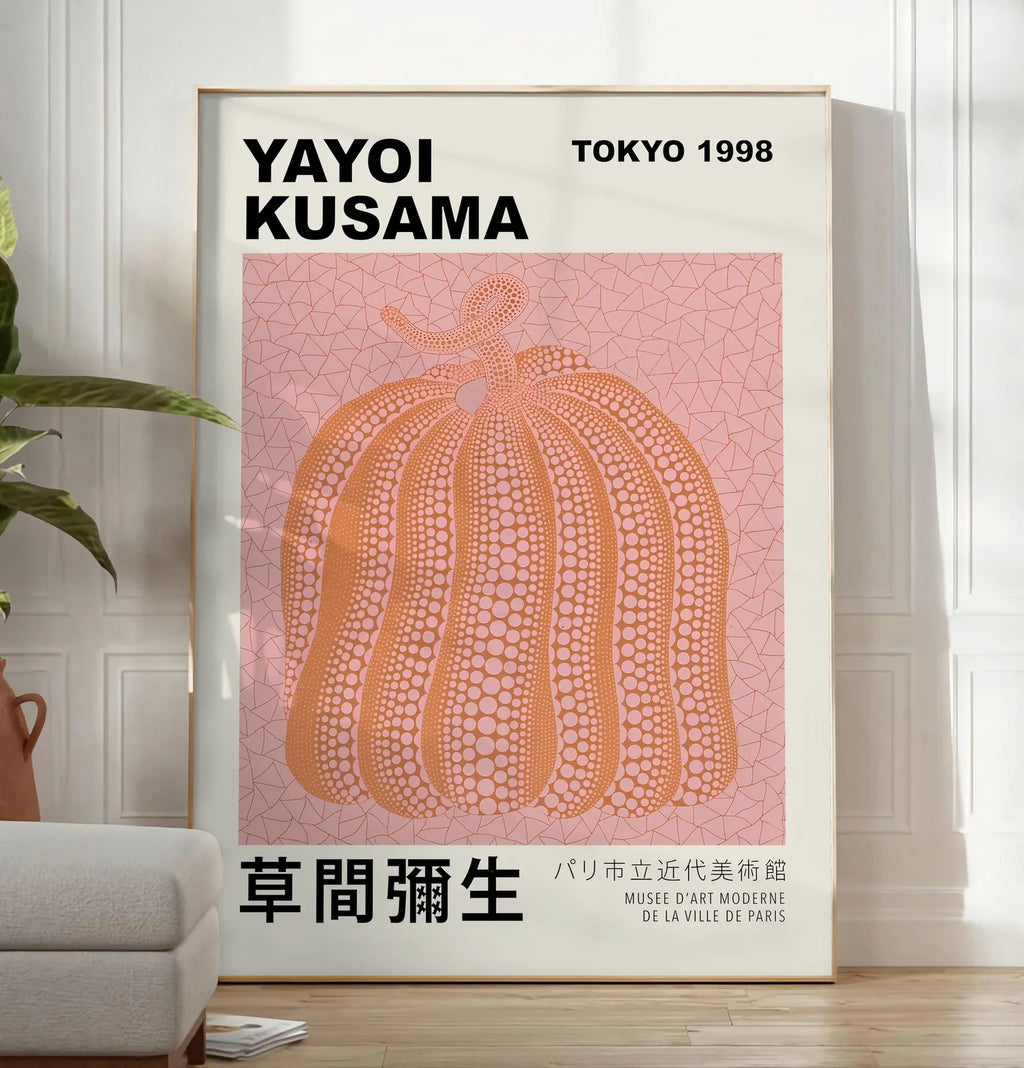 Reproduction affiche Kusama citrouille orange a pois sur fond rose, posee au sol dans un salon minimaliste clair, a cote d'un canape beige et d'une plante verte, sur un parquet clair