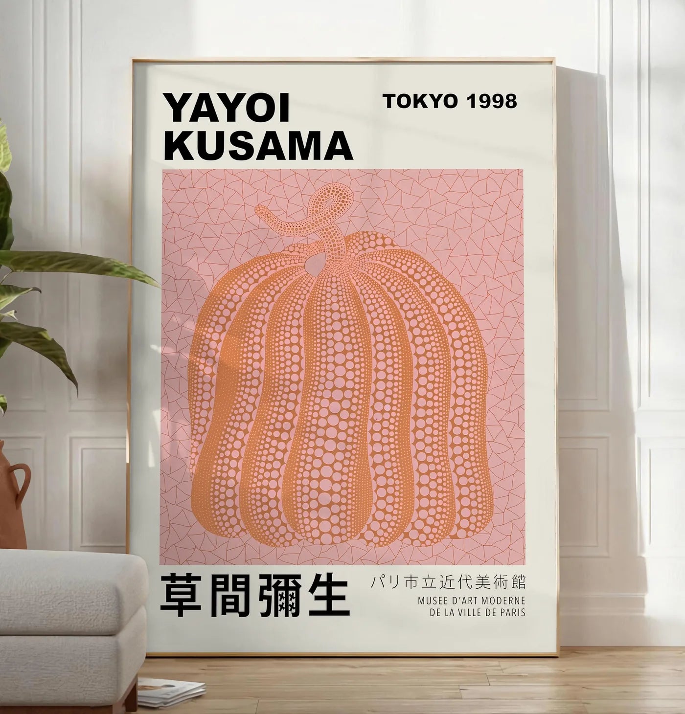 Reproduction affiche Kusama citrouille orange a pois sur fond rose, posee au sol dans un salon minimaliste clair, a cote d'un canape beige et d'une plante verte, sur un parquet clair