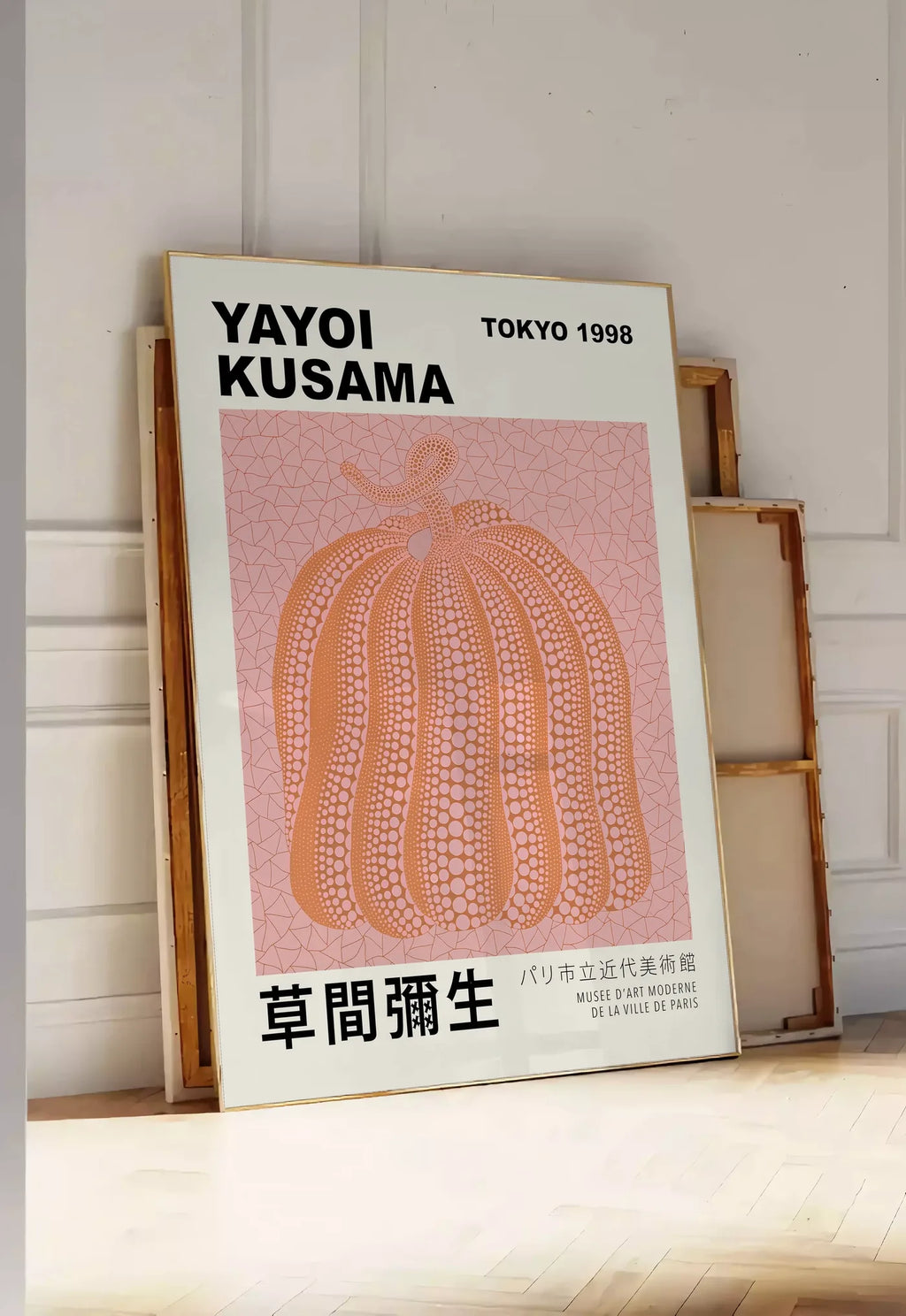 Tirage encadrable affiche Kusama citrouille orange a pois sur fond rose, posee au sol dans une entree lumineuse, contre un mur a panneaux blancs, a cote de chassis en bois empiles