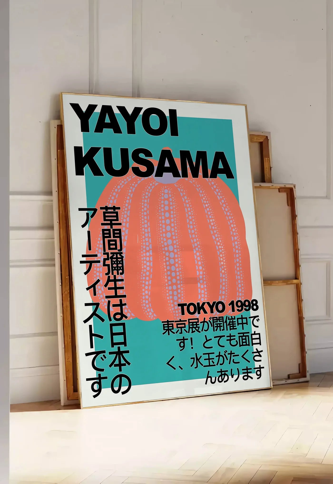 Tirage encadrable affiche citrouille orange de Kusama art japonais, posee au sol dans une entree lumineuse, contre un mur a panneaux blancs, a cote de chassis en bois empiles