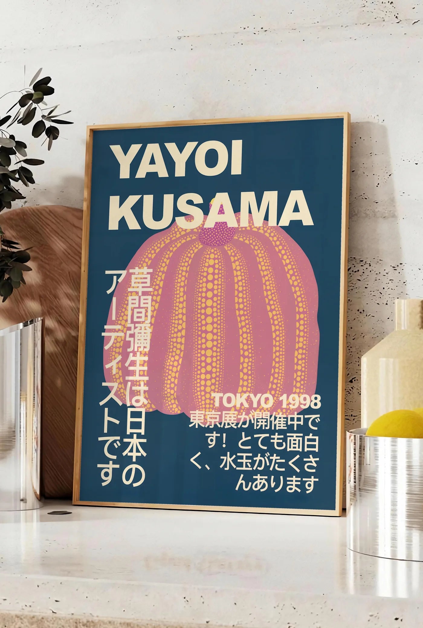 Affiche affiche Kusama citrouille rose a pois sur fond bleu nuit, posee sur un plan de travail de cuisine en pierre claire, entre une branche d'eucalyptus en vase et un bocal de citrons