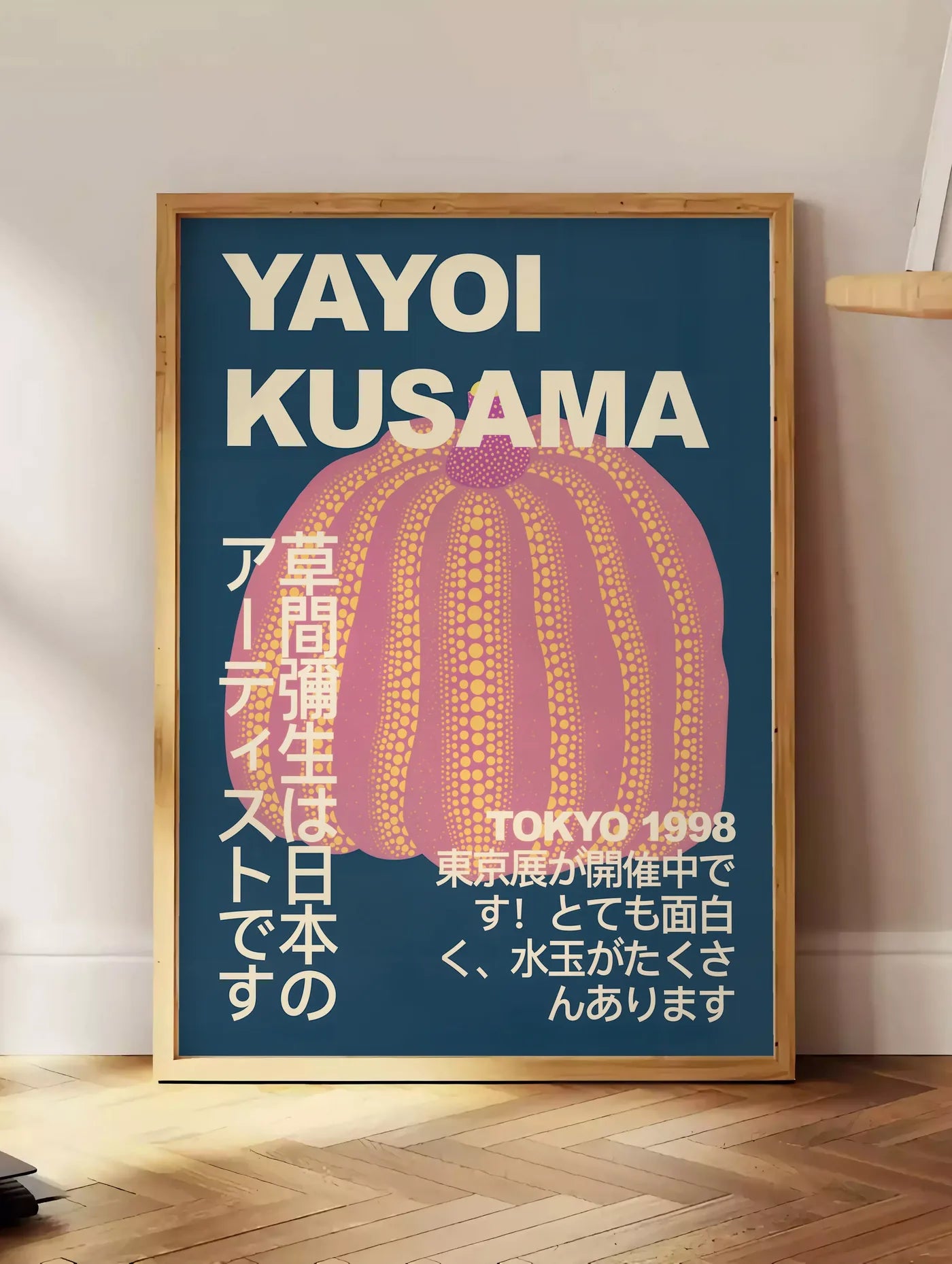 Estampe affiche Kusama citrouille rose a pois sur fond bleu nuit, posee au sol dans un coin salon, sur un parquet a chevrons, pres d'une table d'appoint en chene chargee de livres