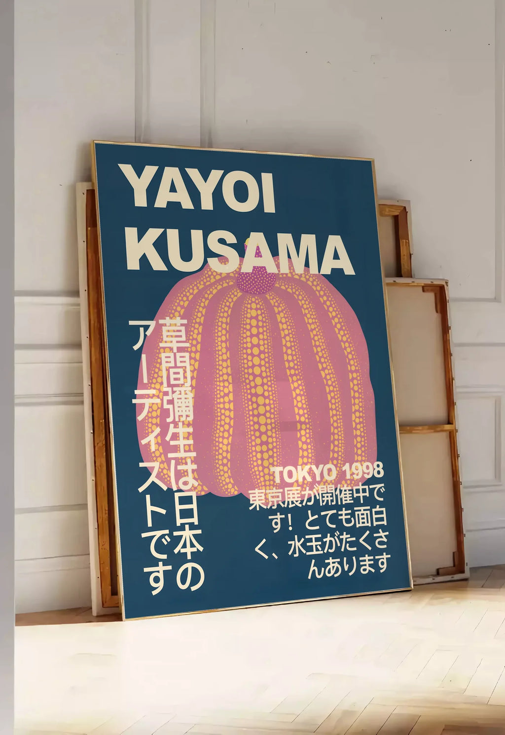 Tirage encadrable affiche Kusama citrouille rose a pois sur fond bleu nuit, posee au sol dans une entree lumineuse, contre un mur a panneaux blancs, a cote de chassis en bois empiles