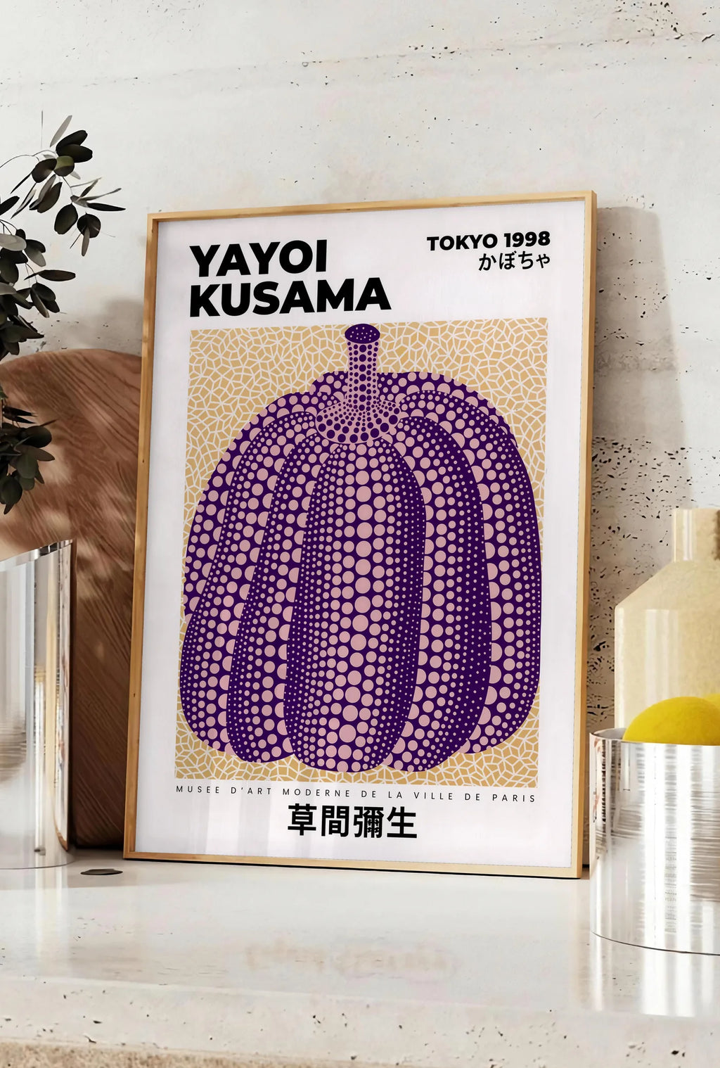 Affiche affiche Kusama citrouille violette a pois sur fond beige, posee sur un plan de travail de cuisine en pierre claire, entre une branche d'eucalyptus en vase et un bocal de citrons