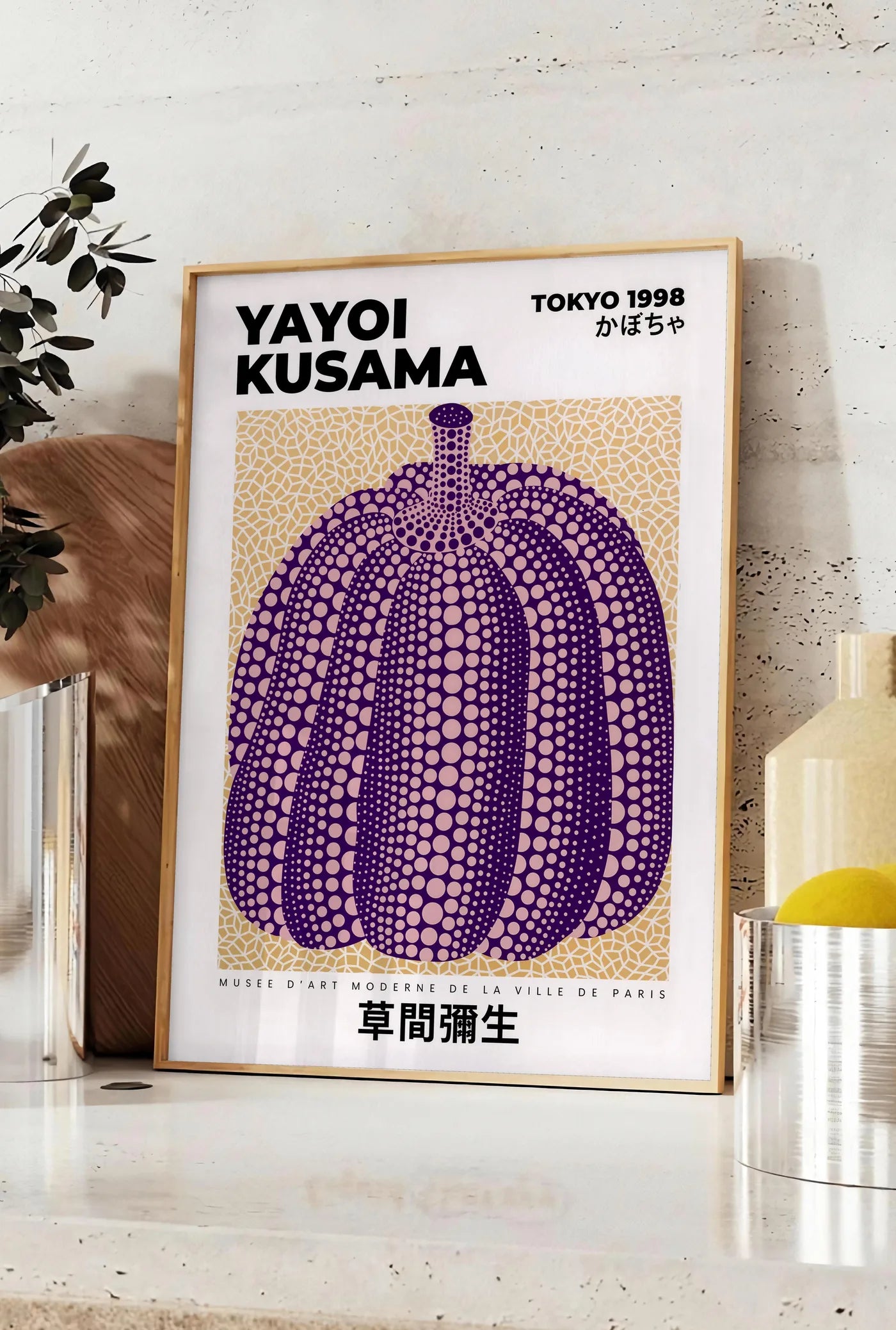 Affiche affiche Kusama citrouille violette a pois sur fond beige, posee sur un plan de travail de cuisine en pierre claire, entre une branche d'eucalyptus en vase et un bocal de citrons