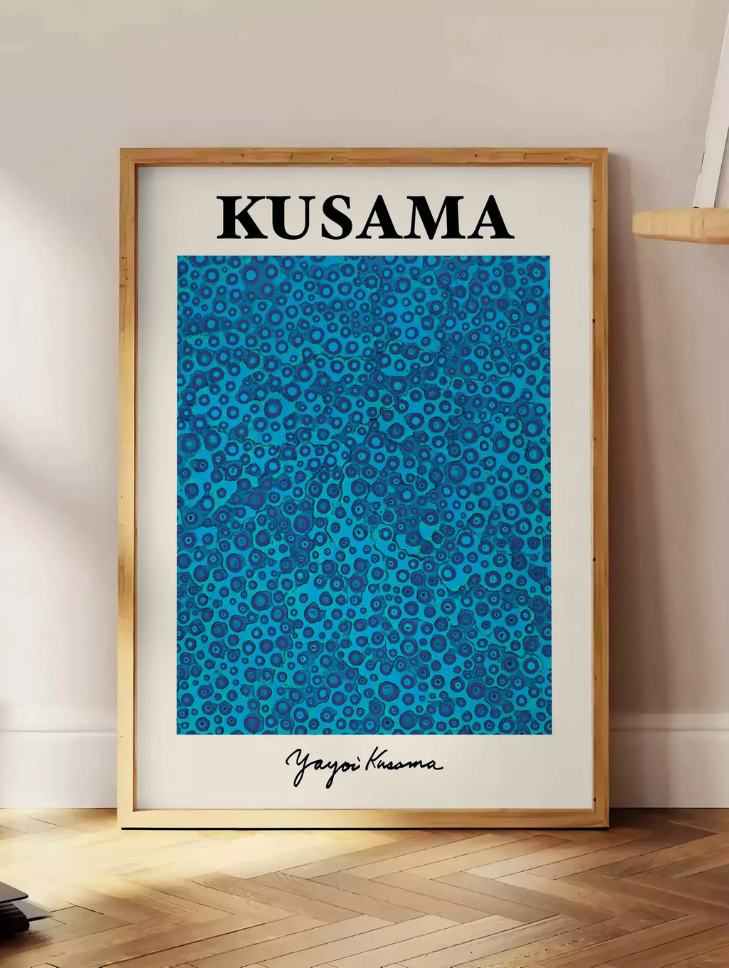 Estampe affiche pois bleus de Kusama art japonais, posee au sol dans un coin salon, sur un parquet a chevrons, pres d'une table d'appoint en chene chargee de livres