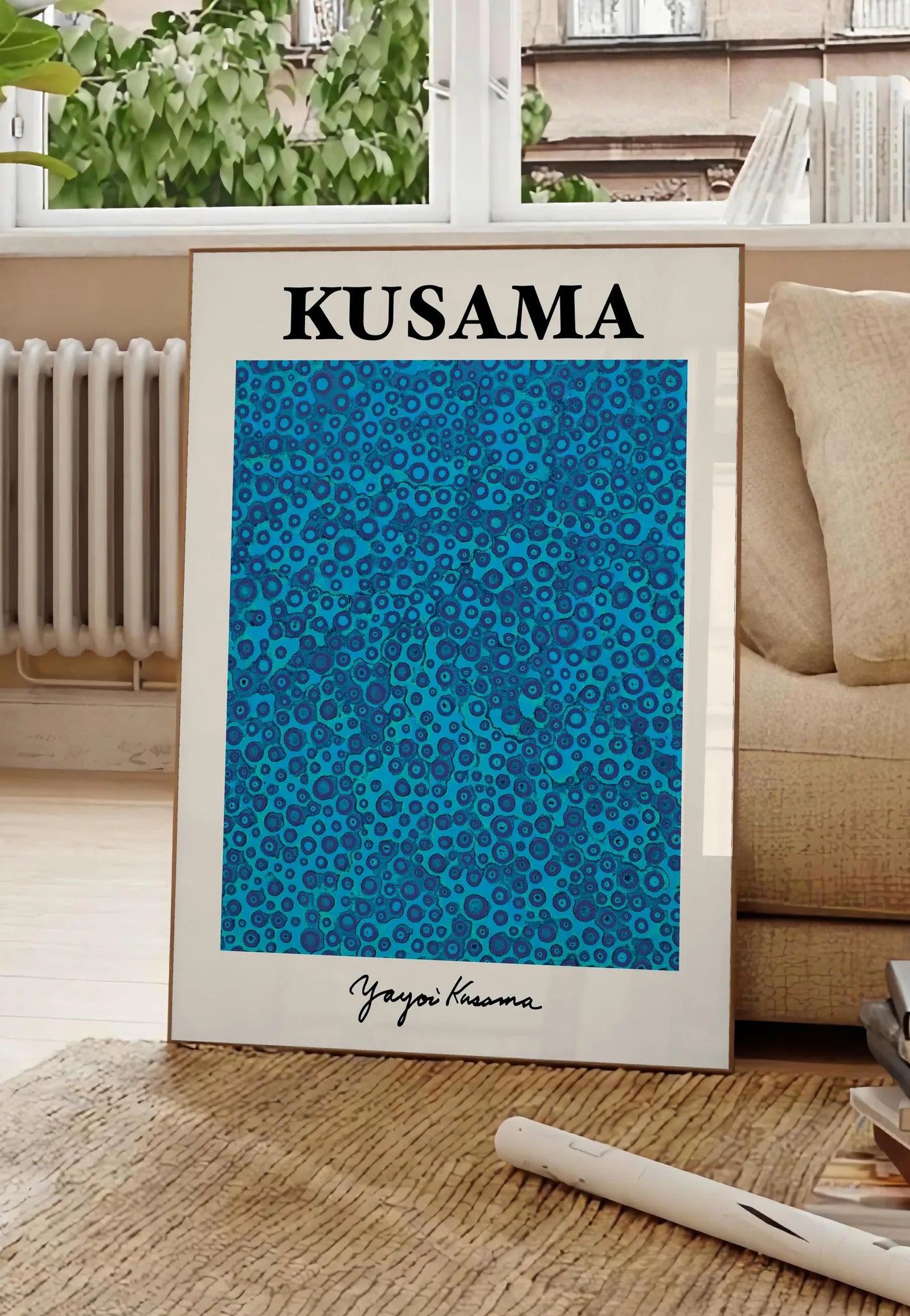 Poster affiche pois bleus de Kusama art japonais, posee au sol dans un salon parisien, devant une grande fenetre donnant sur un immeuble haussmannien, entre un ficus et un canape creme