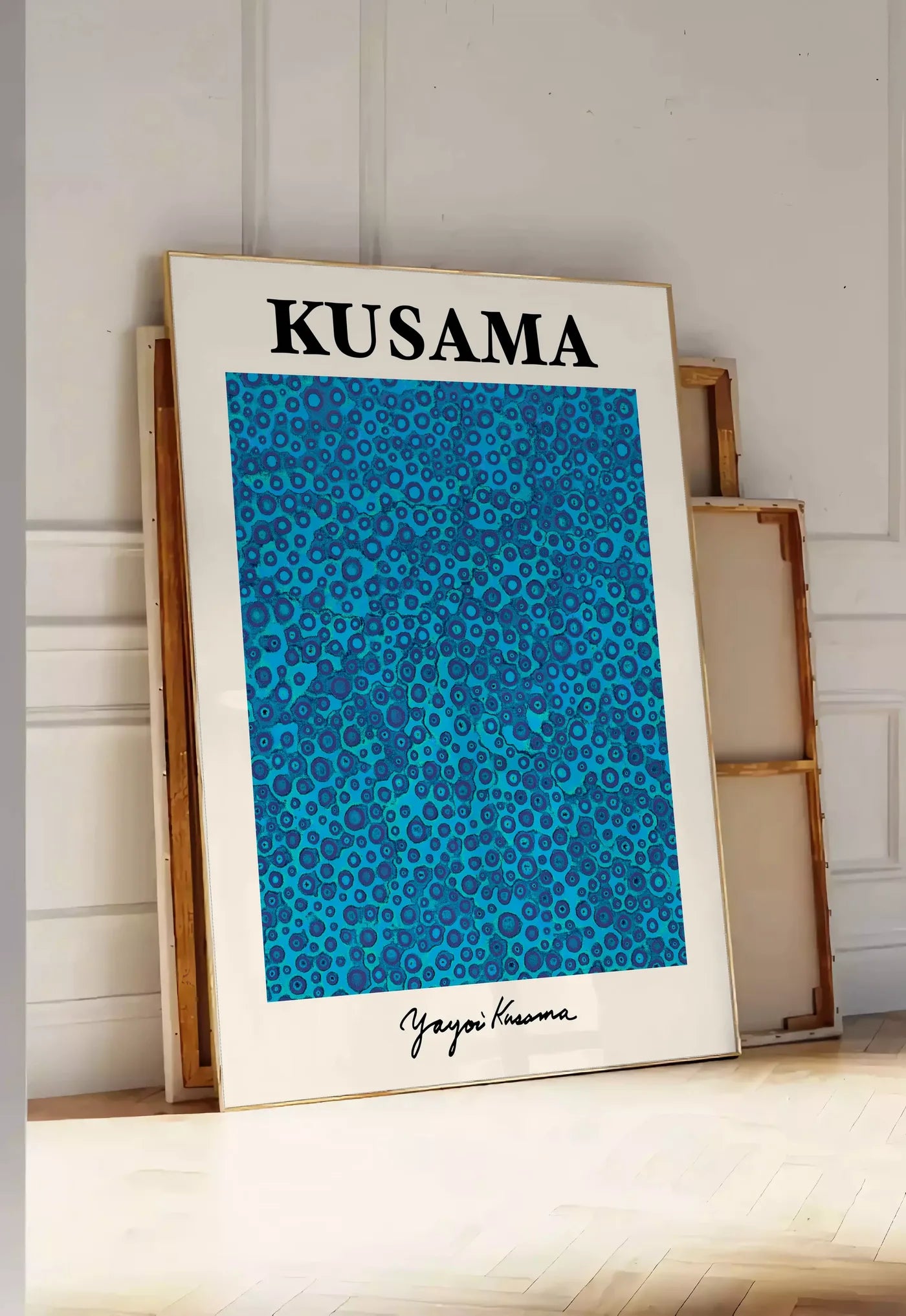 Tirage encadrable affiche pois bleus de Kusama art japonais, posee au sol dans une entree lumineuse, contre un mur a panneaux blancs, a cote de chassis en bois empiles